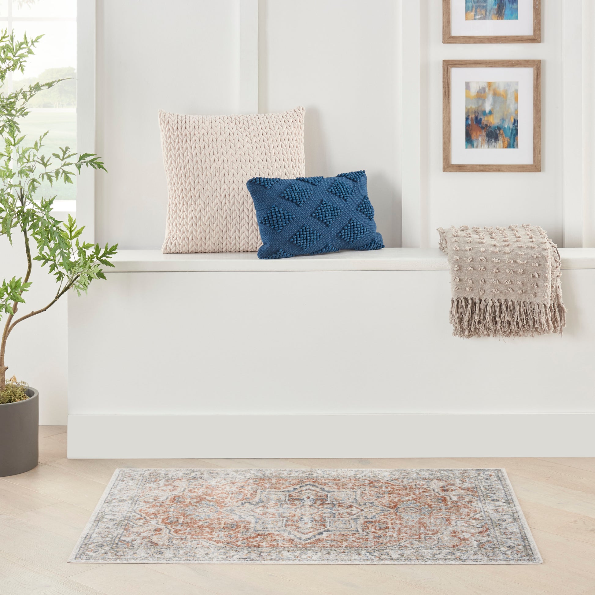 Bright living nook with a beige knitted cushion, a blue textured pillow, taupe fringe throw, plant, framed art, and a patterned rug.