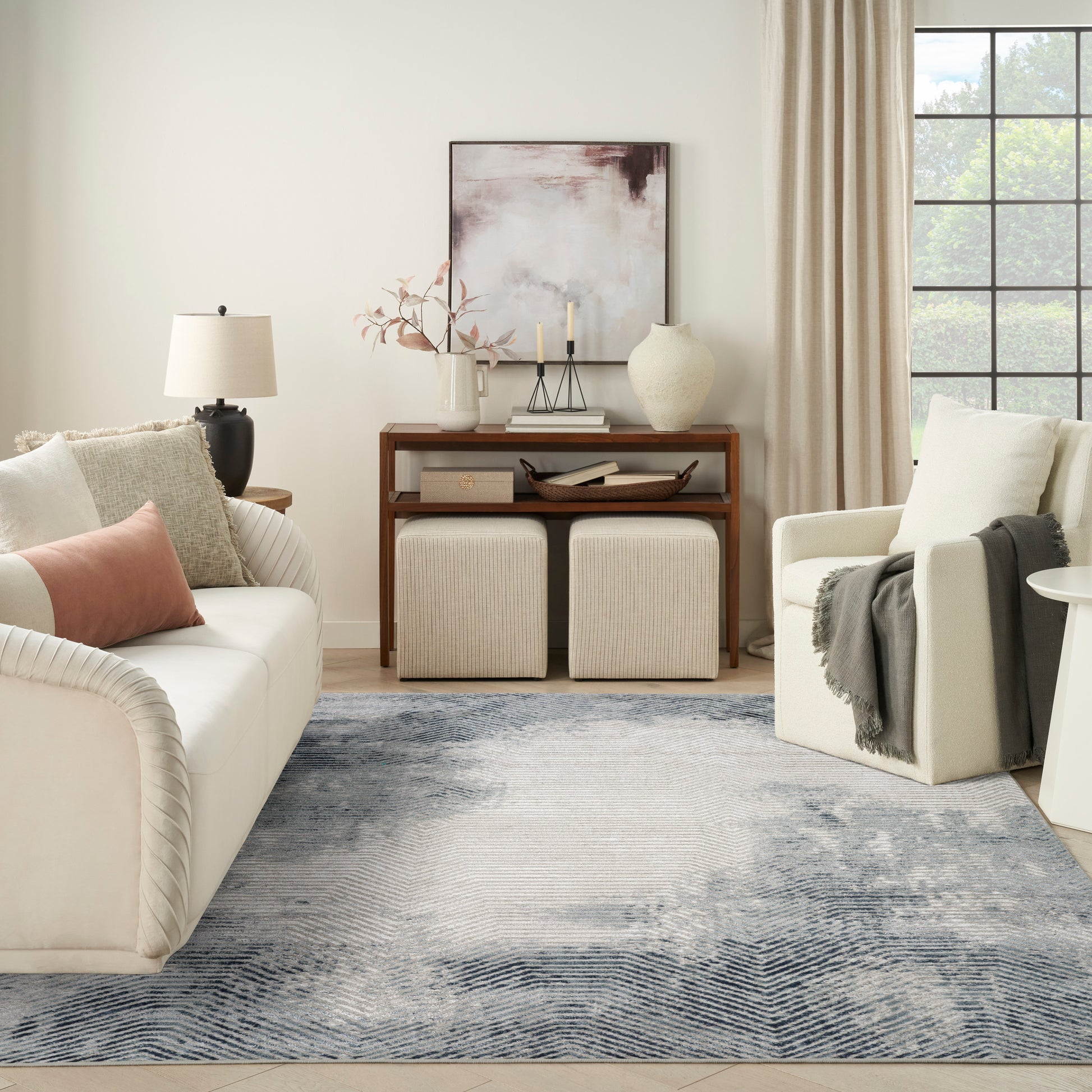 Neutral living room with cream sofa, blush pillow, wooden console, ottomans, abstract art, and a blue-gray rug.