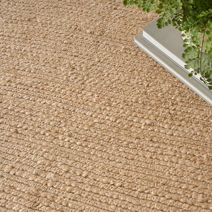 Natural beige jute rug with thick loop texture; green fern and silver-edged decor at the top-right.