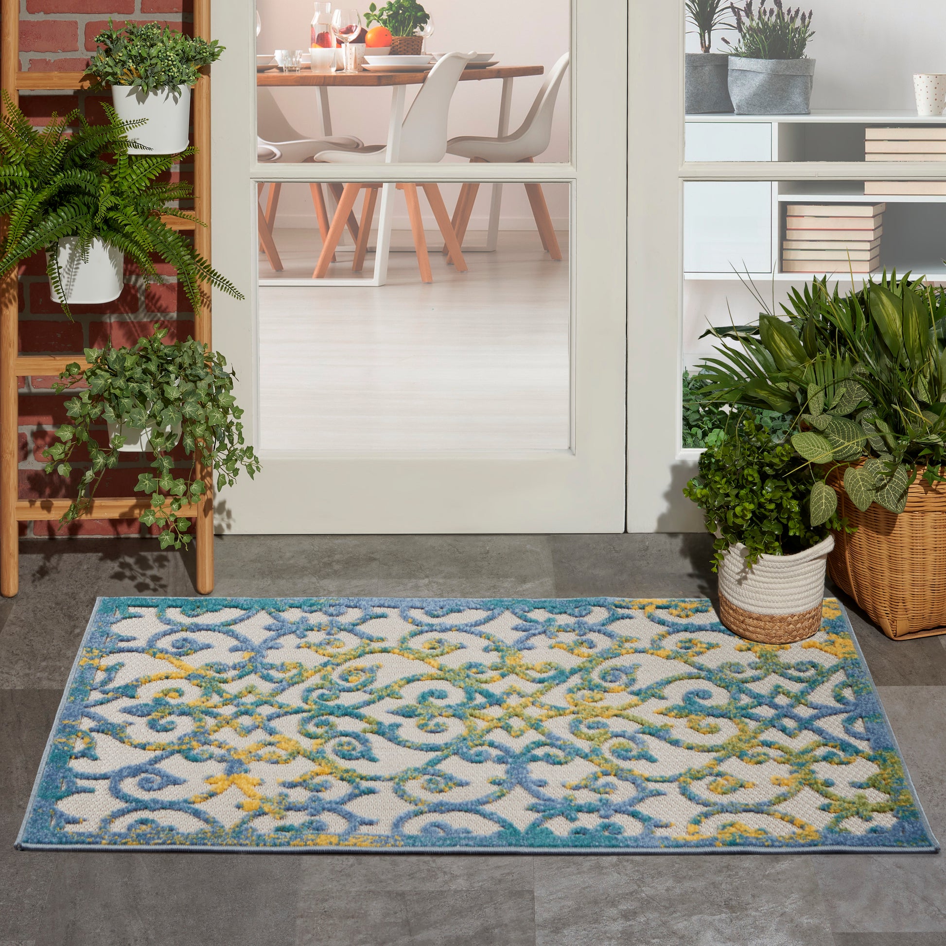 Outdoor doormat with blue teal scroll pattern and yellow accents in front of a glass door; potted ferns nearby.