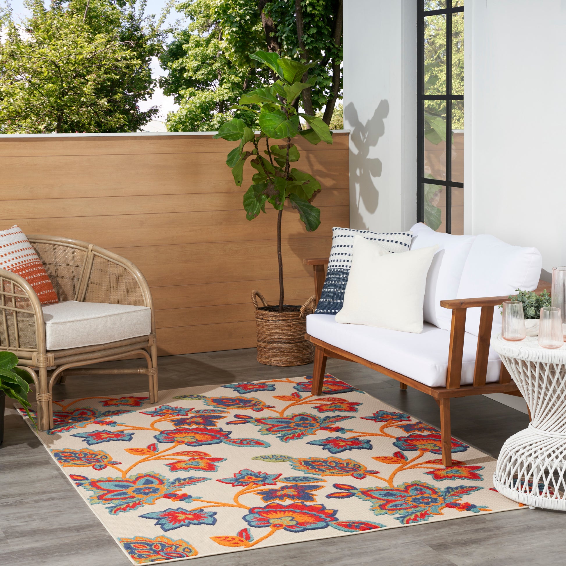 Cozy outdoor patio with white cushioned sofa, wicker chair, vibrant floral rug, potted plant, wooden screen and glass door.