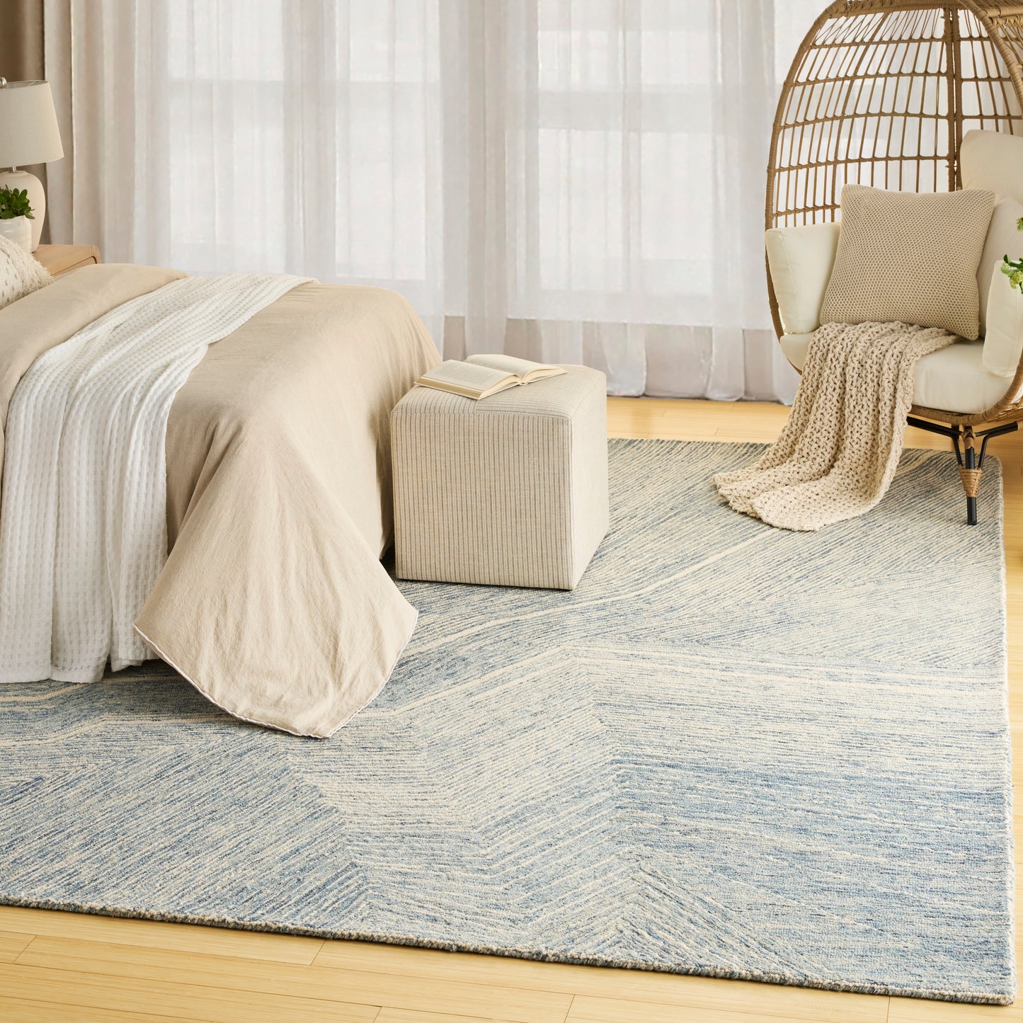 Neutral bedroom with beige bedding, blue-gray rug, cream ottoman, wicker hanging chair, knitted throw, sheer curtains.