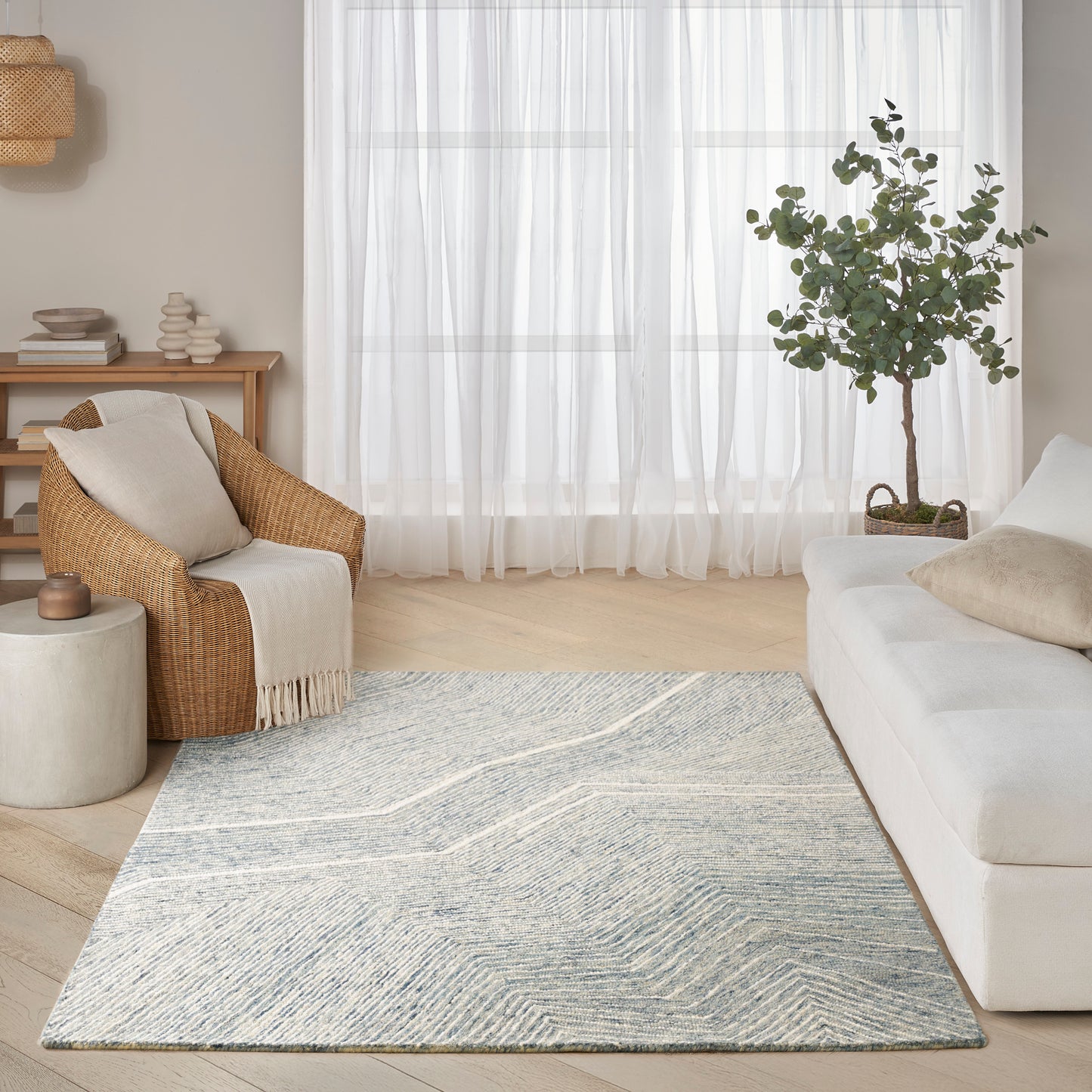 Rectangular blue-gray geometric rug in a light modern living room with white sofa, wicker armchair, and sheer curtains.