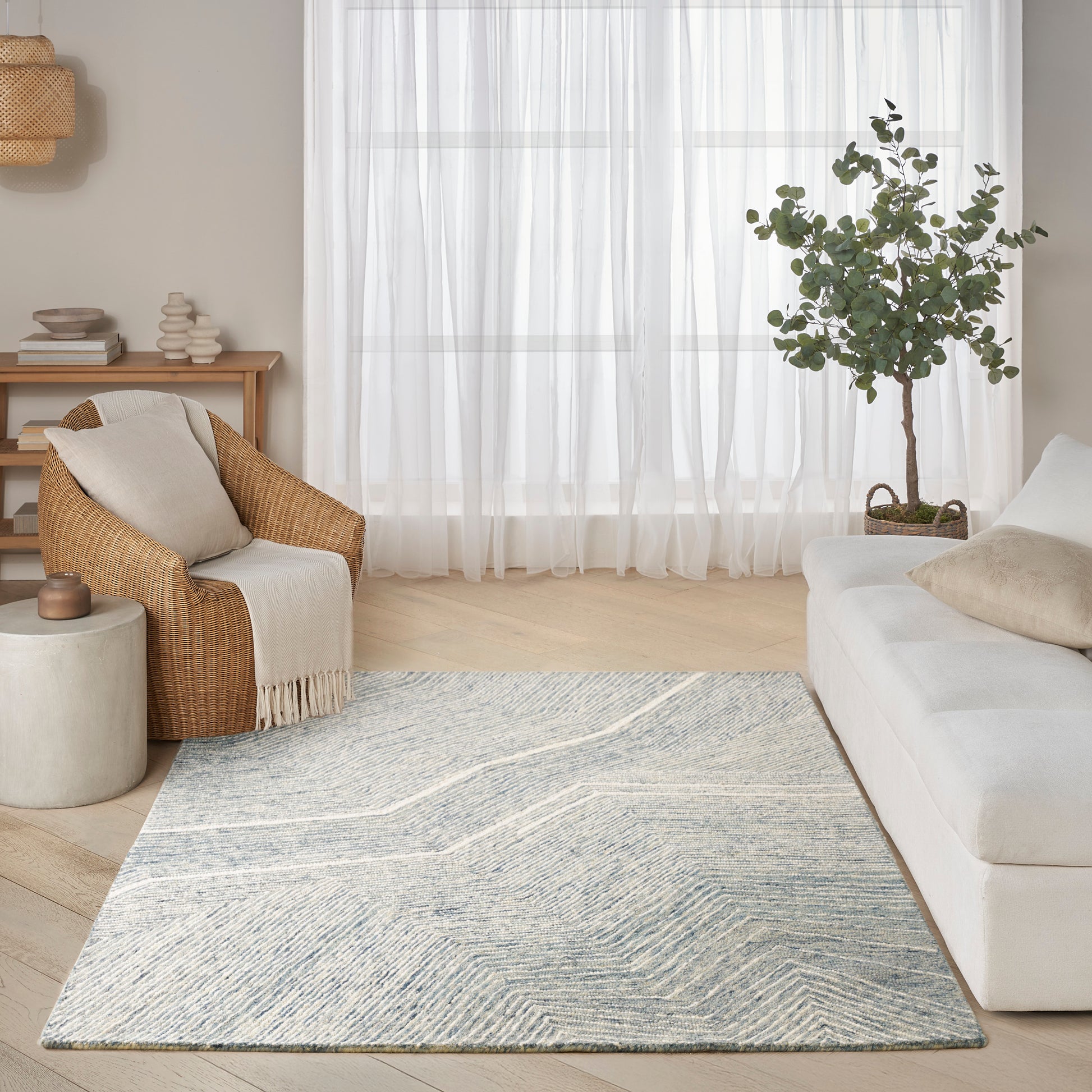 Rectangular blue-gray geometric rug in a light modern living room with white sofa, wicker armchair, and sheer curtains.