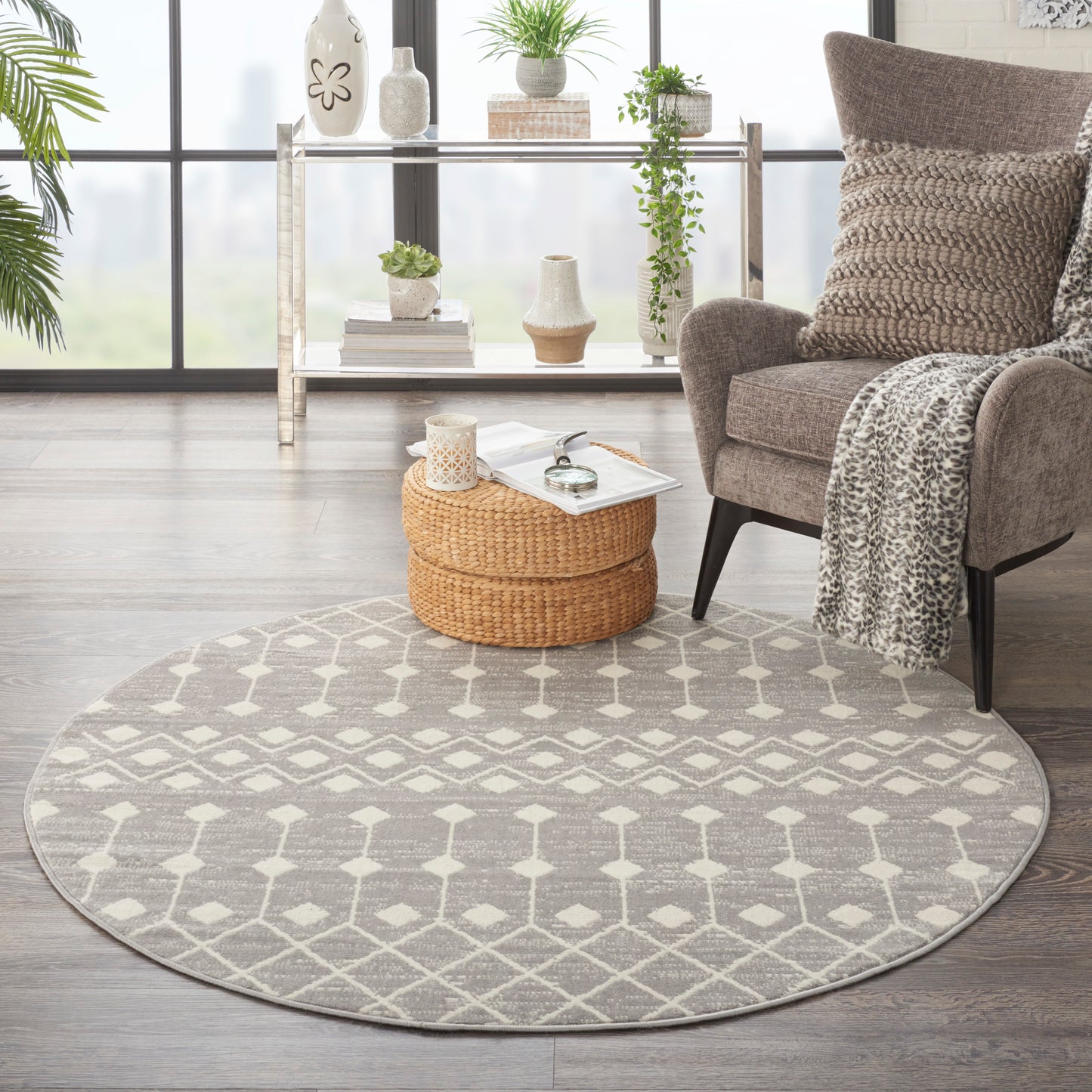 Round gray rug with white lattice pattern in a modern living room; woven ottoman, beige armchair, knit throw, plants.