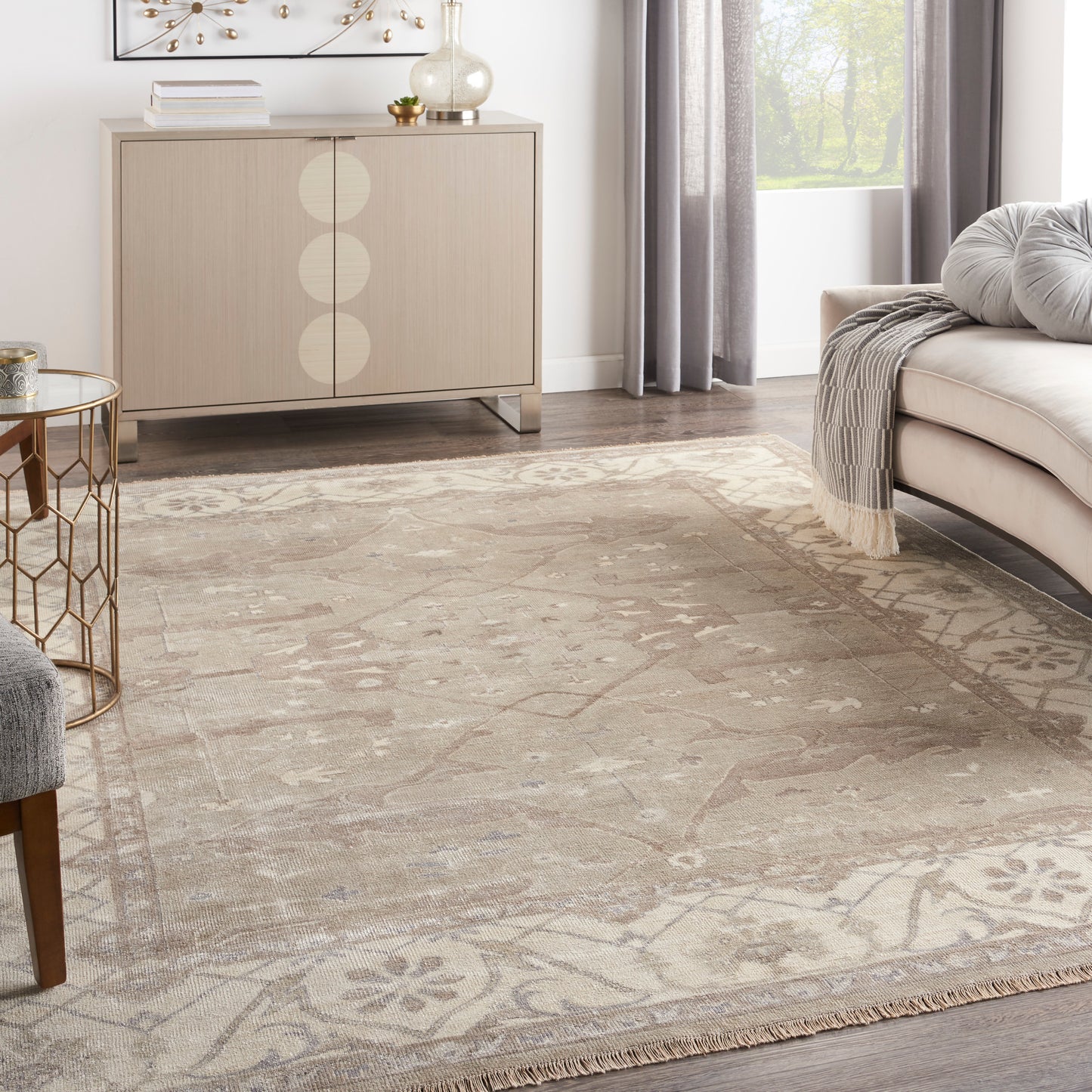 Neutral living room with rug, light wood sideboard with circles, gray sofa, glass-top gold side table, gray curtains.