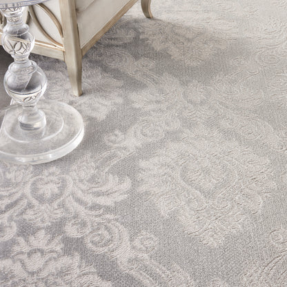 Light gray damask-pattern rug with floral motifs beside a clear glass table and beige sofa.
