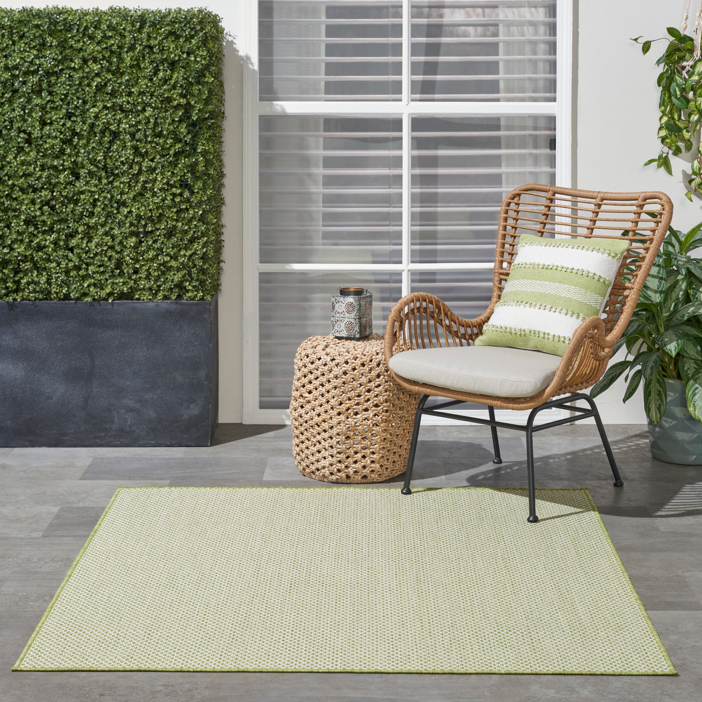 Outdoor rug in light green, 4' x 4' square, under wicker seating with cream cushions and a green striped pillow beside a woven round side table.