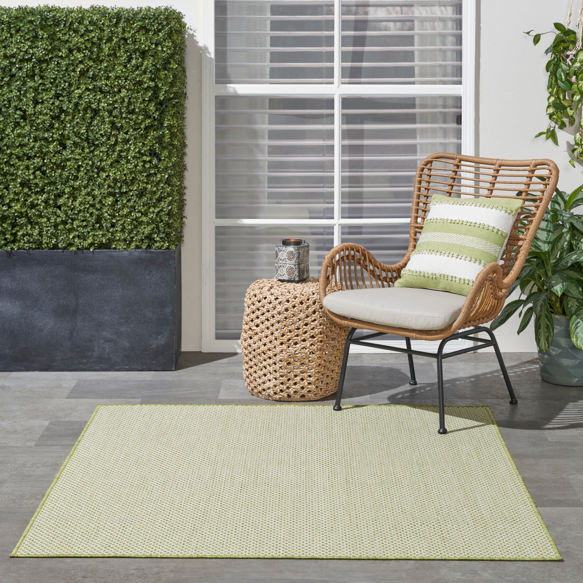 Outdoor rug in light green, 4' x 4' square, under wicker seating with cream cushions and a green striped pillow beside a woven round side table.