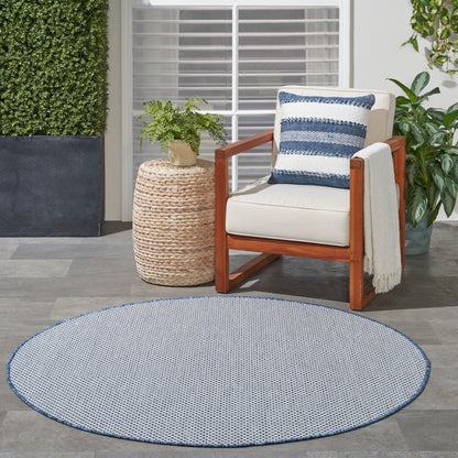 Round blue-and-white outdoor rug with a cream-cushion wooden chair, navy striped pillow, and woven side table with a fern.