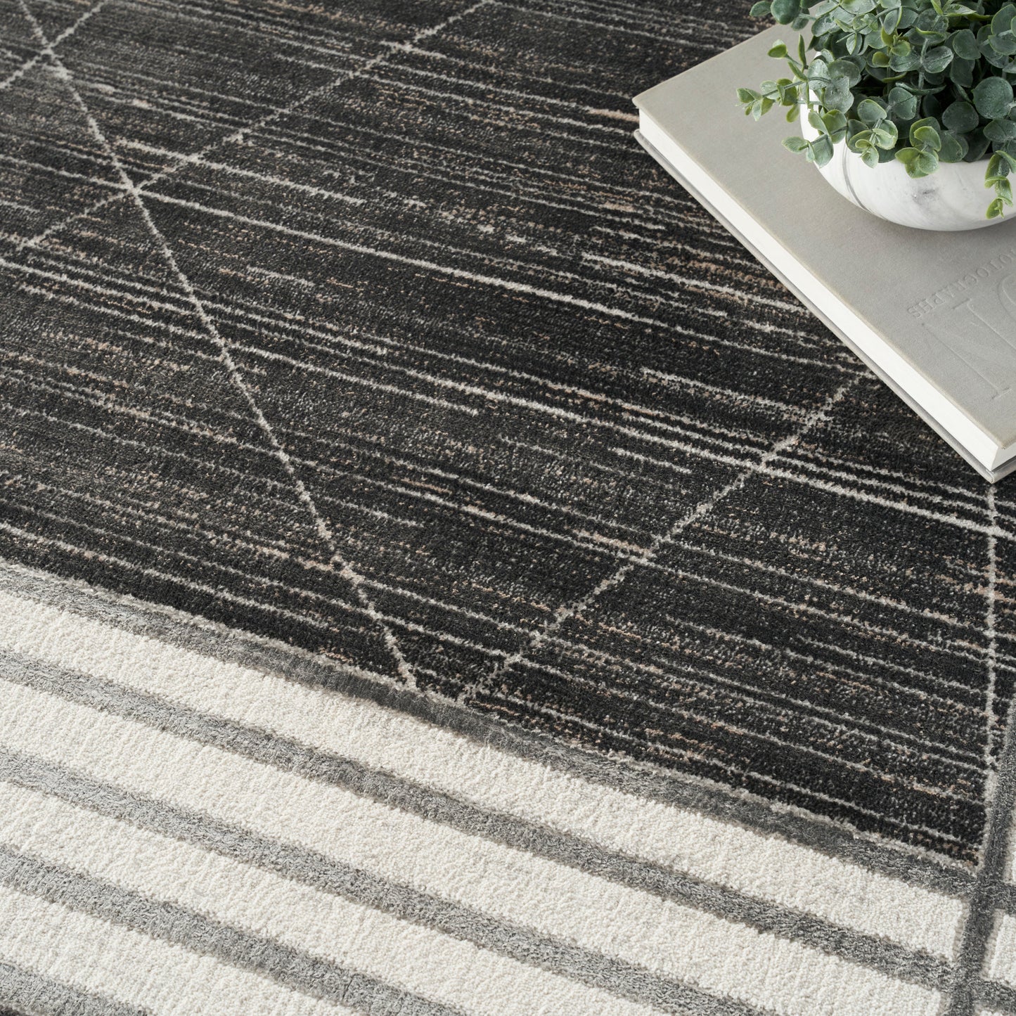 Charcoal rug with white diagonal grid pattern and cream striped border, paired with books and a potted plant.