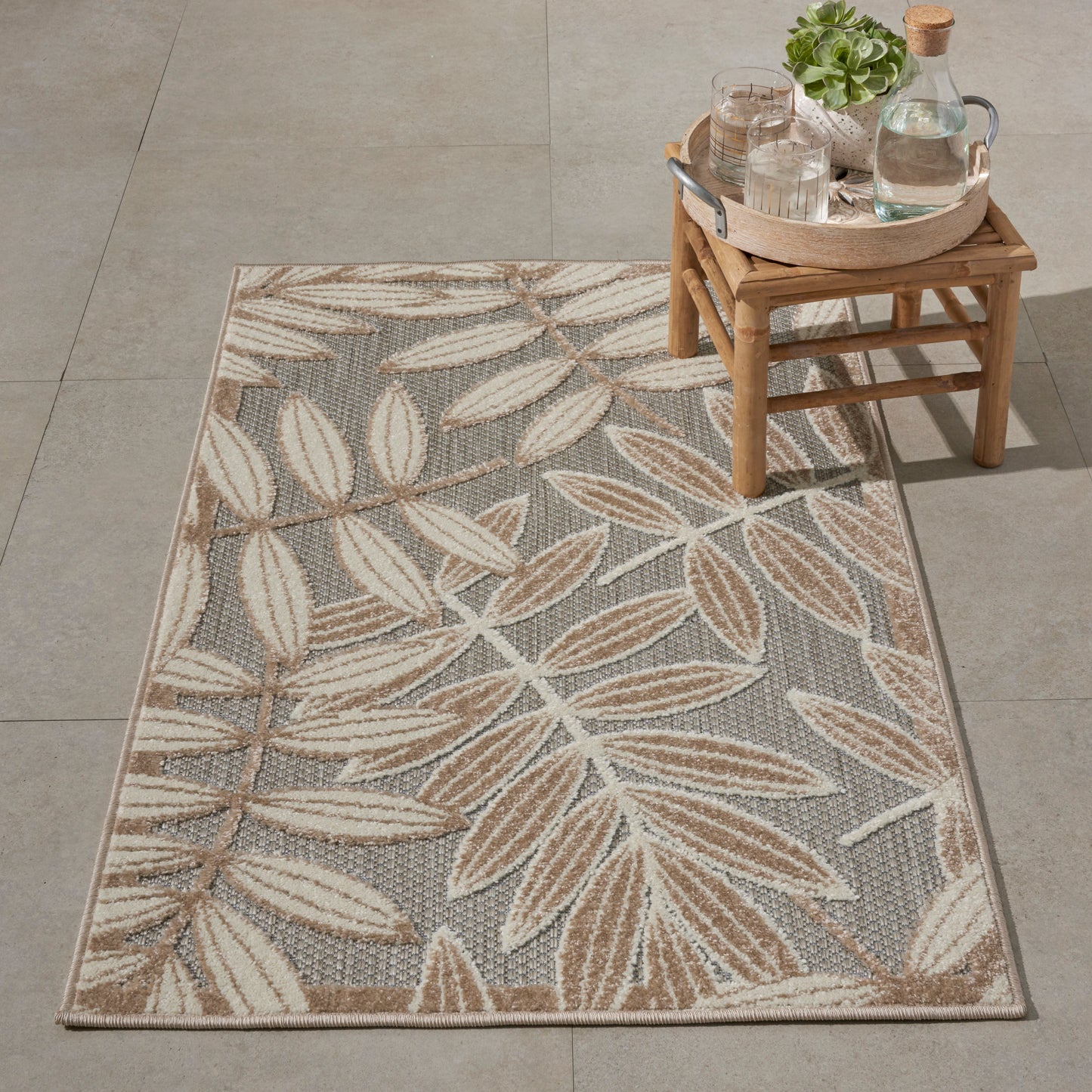 Gray rectangular rug with beige leaf pattern on tiled floor; wooden stool with round tray of glassware, bottle and plant.