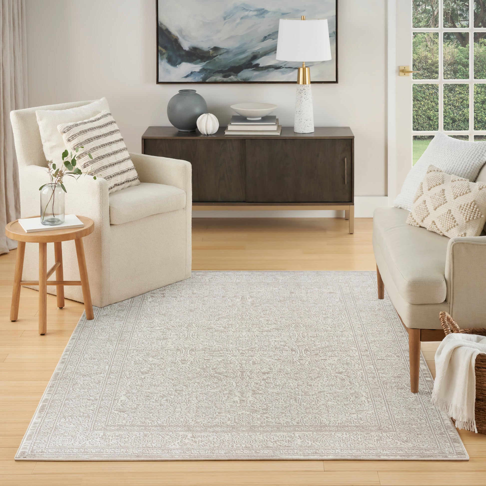 Light gray patterned area rug in a cream living room with beige sofa, armchair, round wood side table, and dark sideboard.
