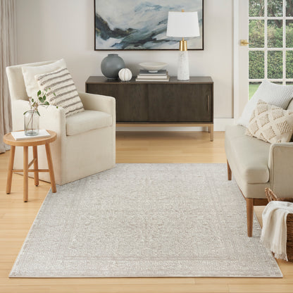 Light gray patterned area rug in a cream living room with beige sofa, armchair, round wood side table, and dark sideboard.