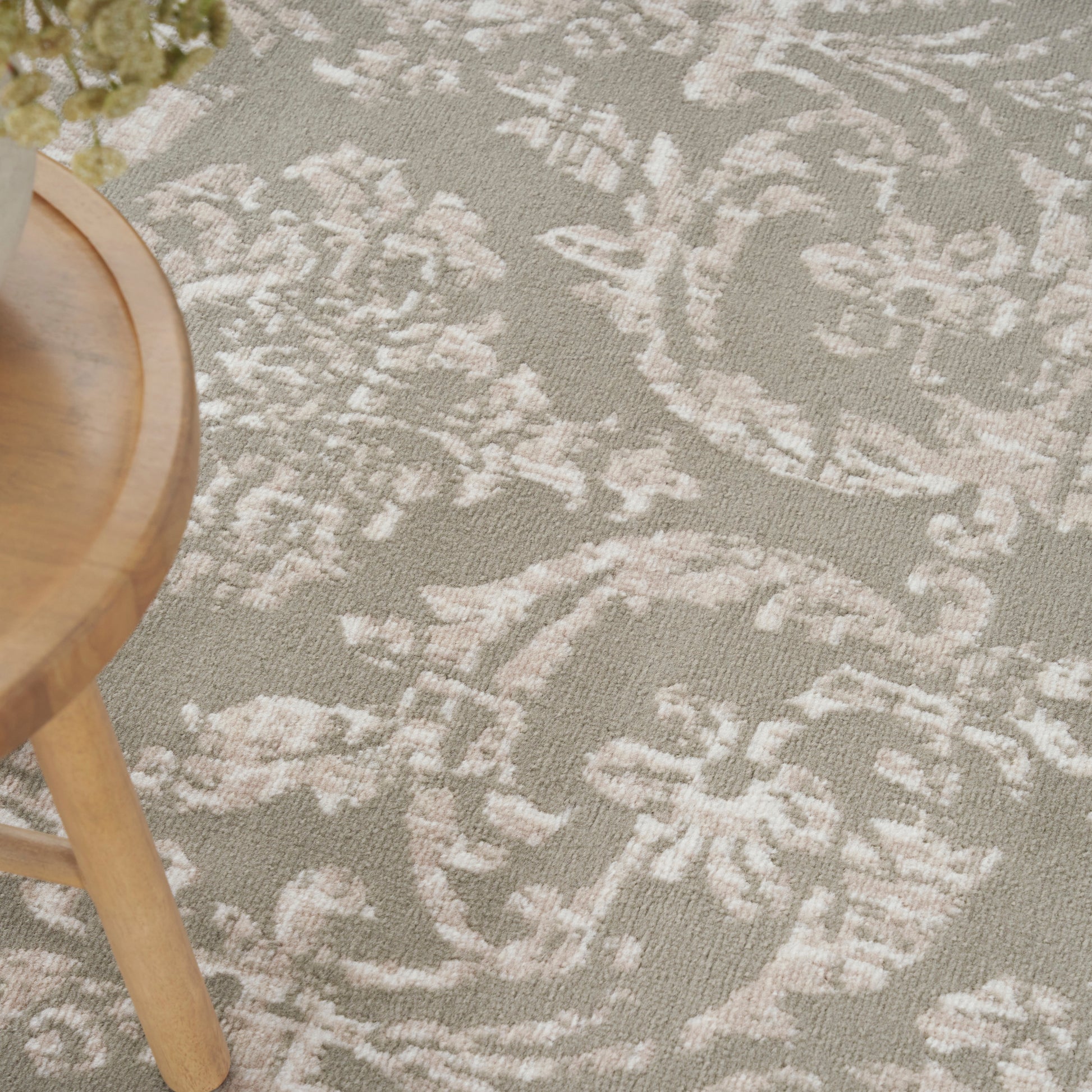 Woven rug with ivory damask floral pattern on taupe beige background & light wood round stool visible.