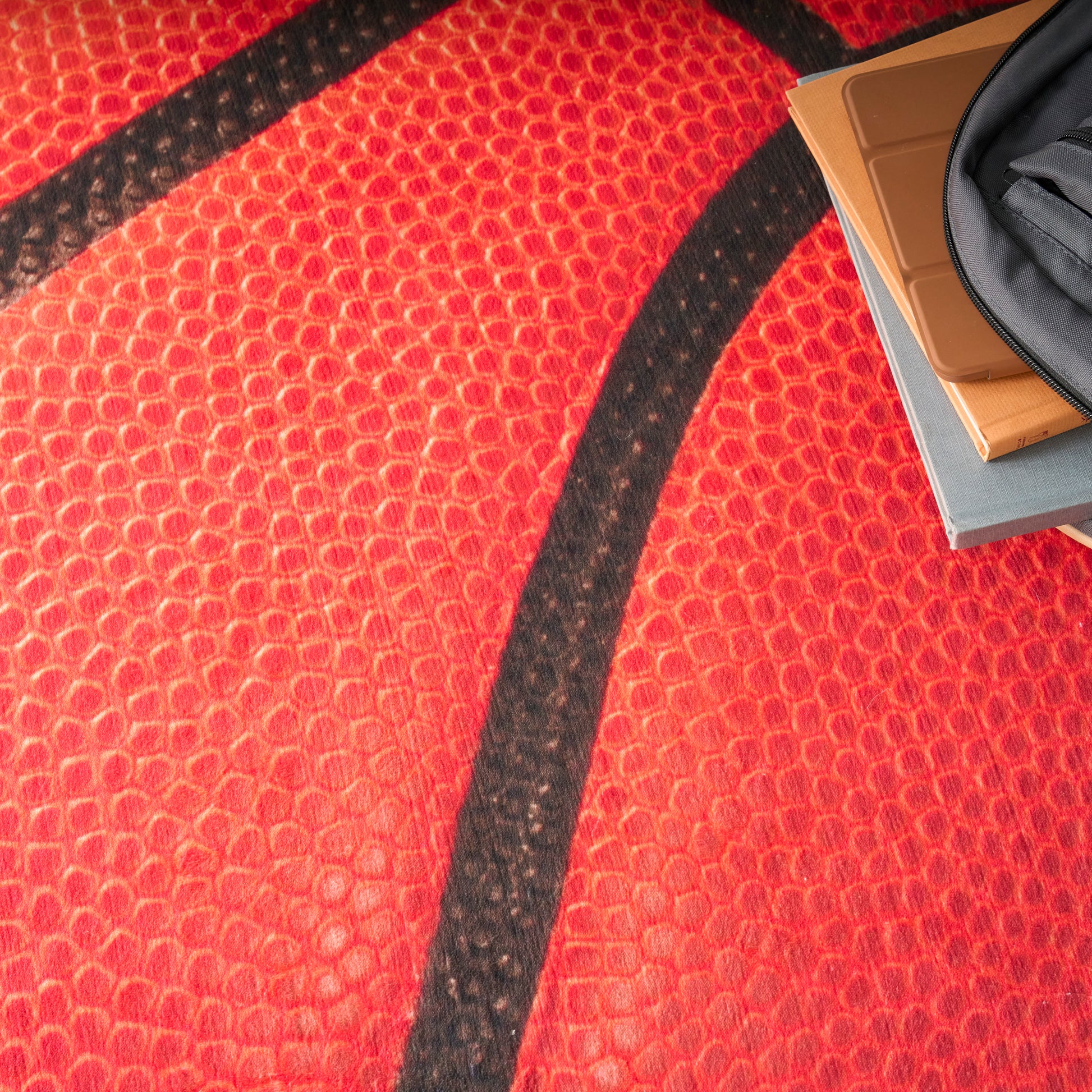 Close-up of red basketball texture with black seam; corner shows a gray zip pouch and orange notebooks.