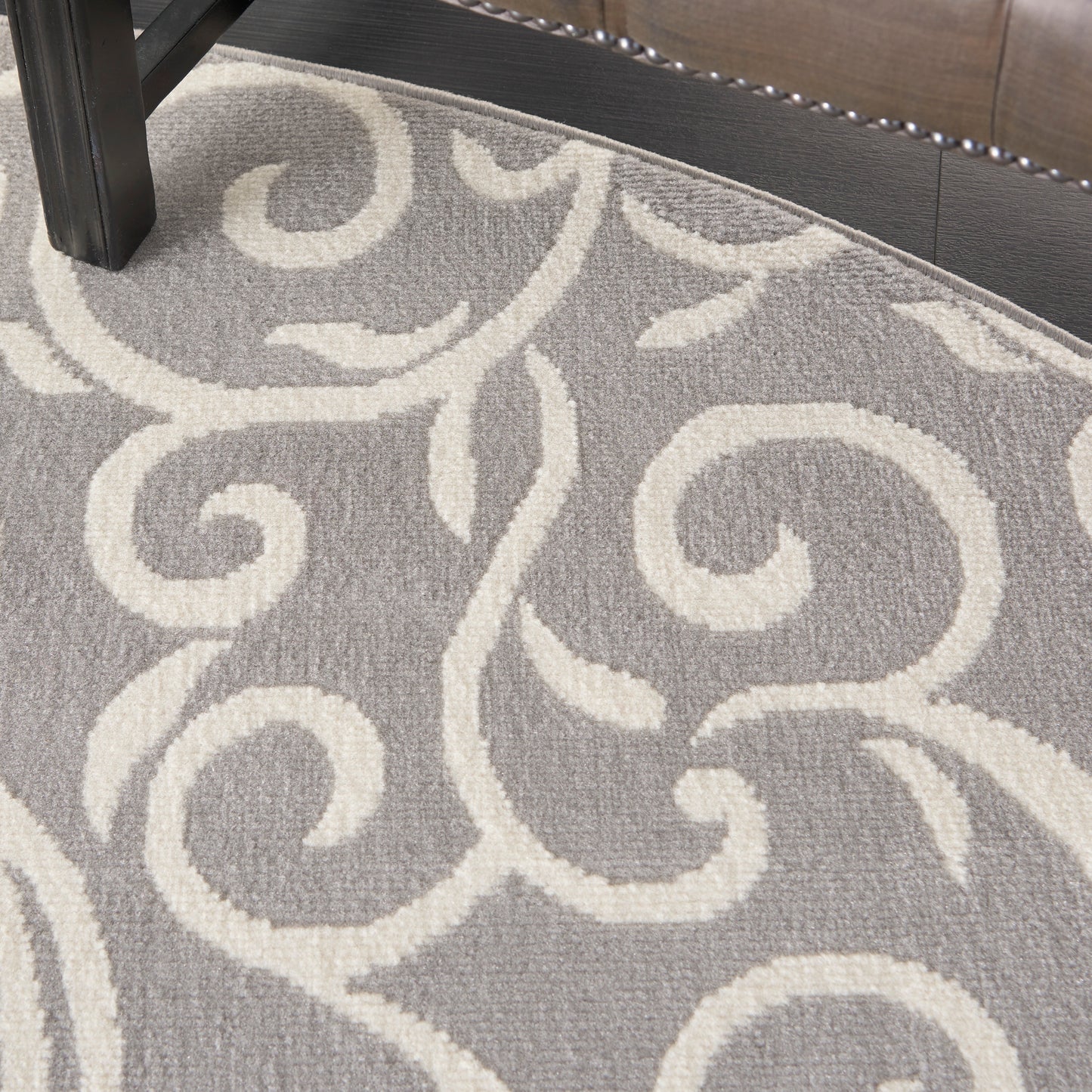 Gray round area rug with ivory scrolling vine motif; plush pile, under a dark wood furniture leg.