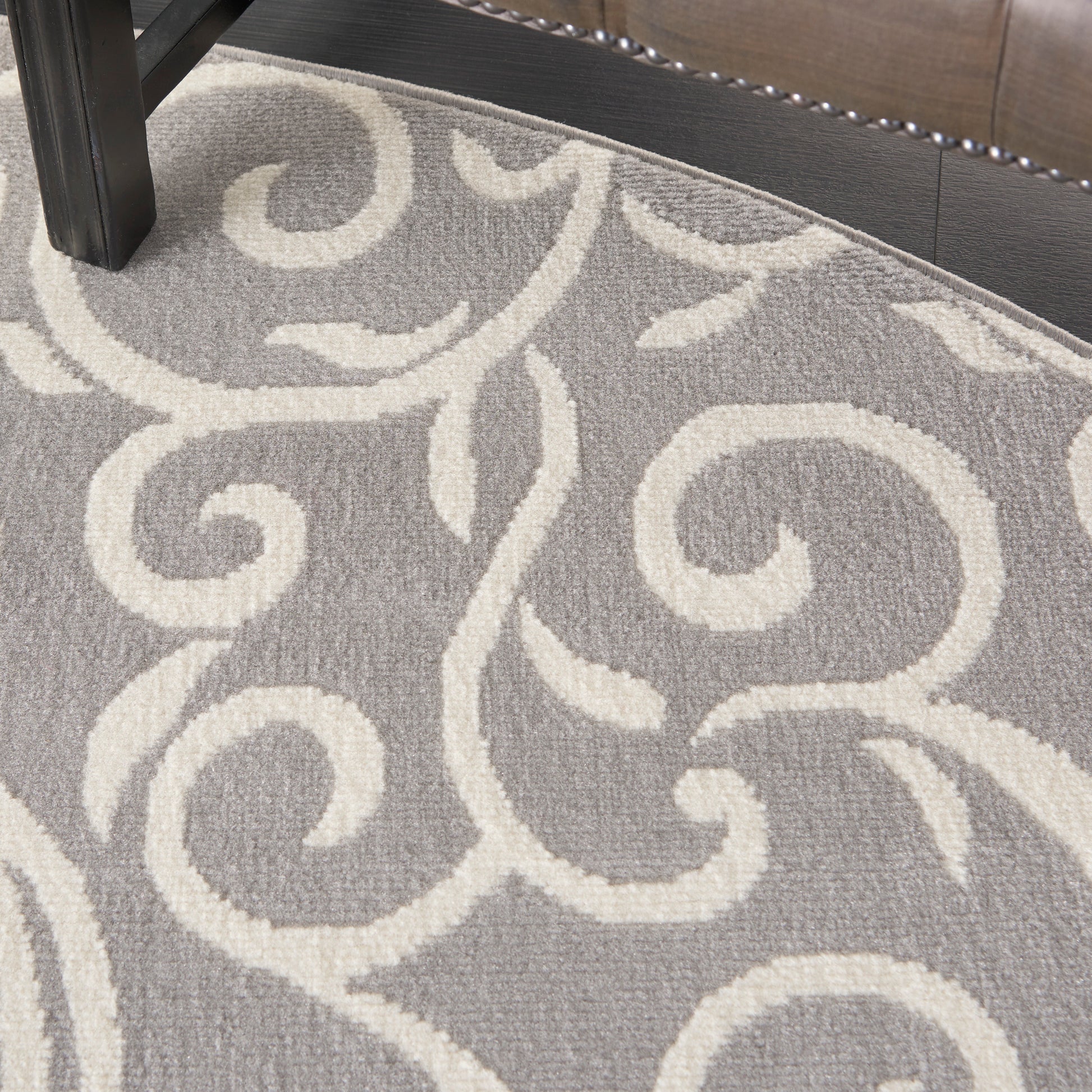 Gray round area rug with ivory scrolling vine motif; plush pile, under a dark wood furniture leg.