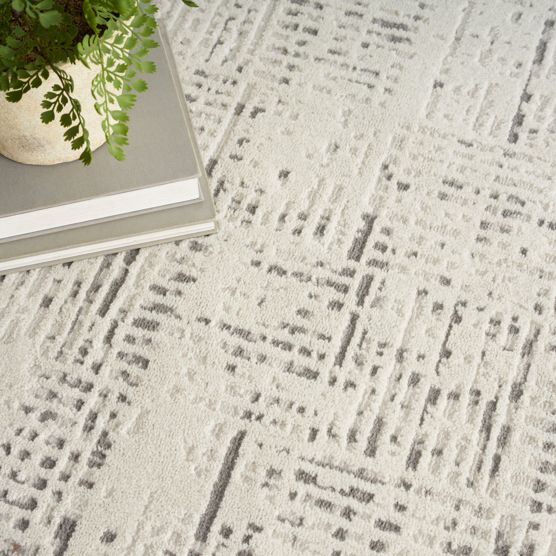 Light gray round area rug with a textured geometric pattern; beige pot and fern sit atop a stack of gray books.
