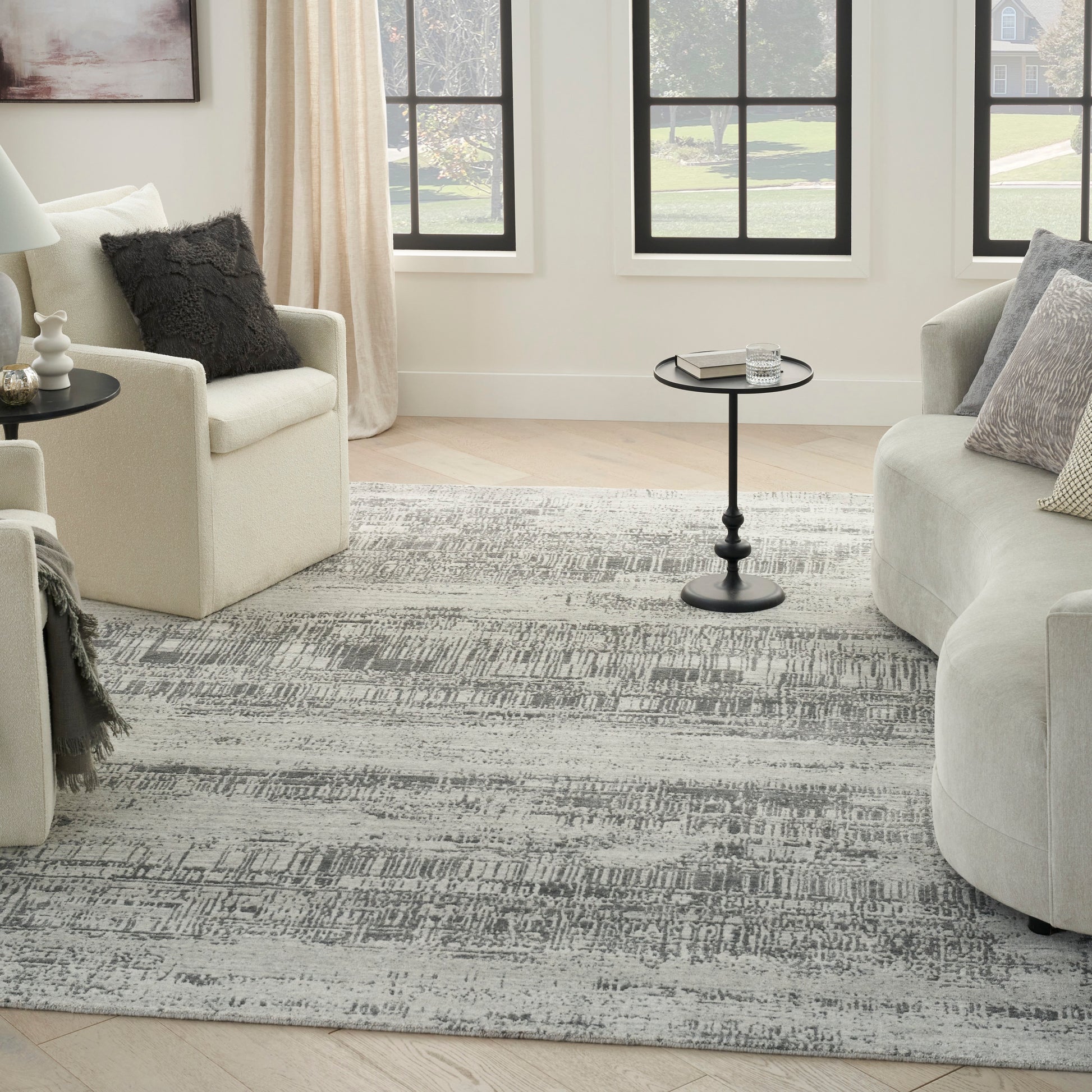 Bright living room with cream armchairs, gray rug, round black pedestal tables, beige curtains, black-framed windows.