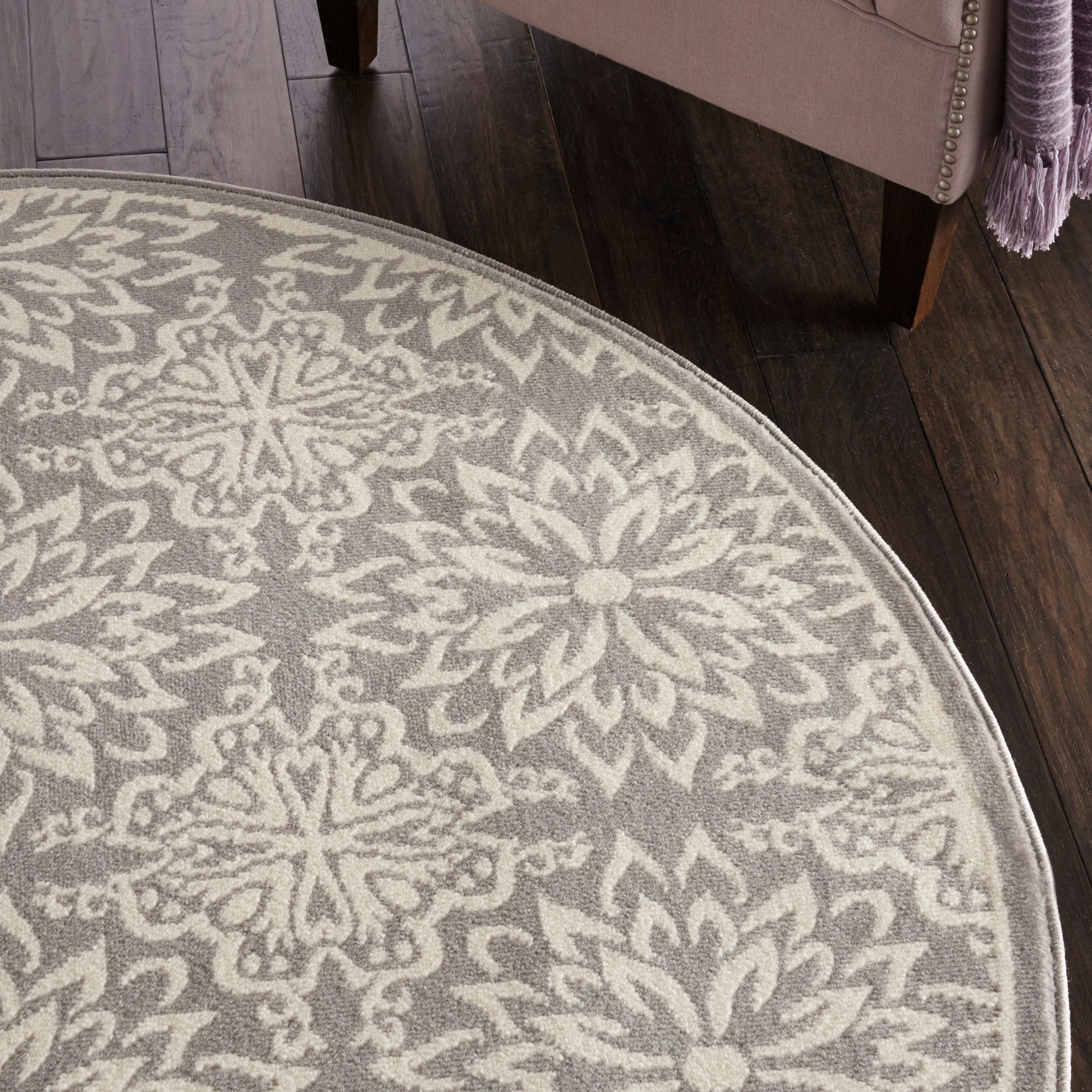 Round grey rug with cream floral medallion pattern; soft plush pile on dark wood floor beside a pink upholstered chair.
