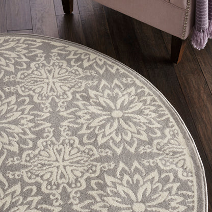 Round grey rug with cream floral medallion pattern; soft plush pile on dark wood floor beside a pink upholstered chair.