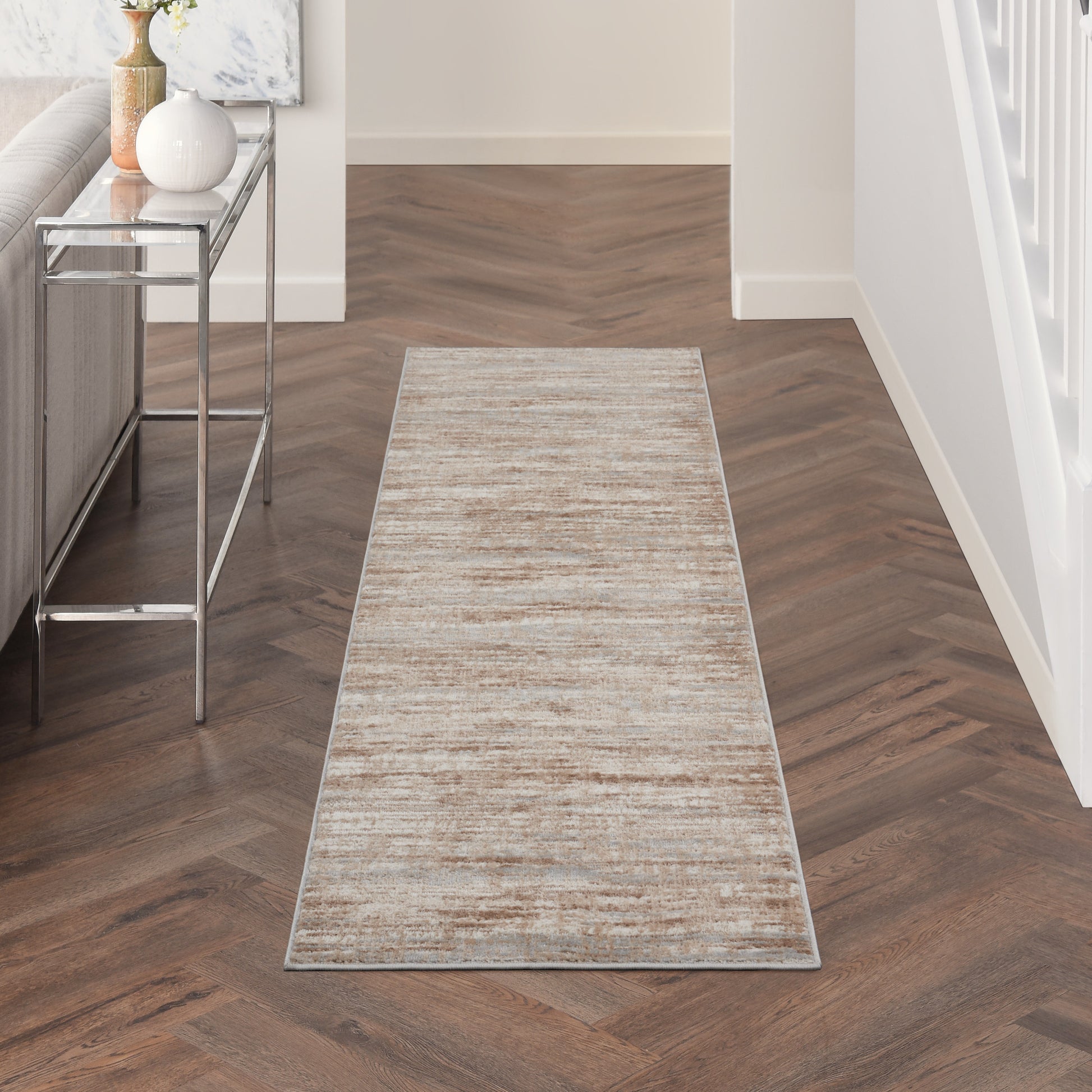 Beige textured hallway runner rug on dark herringbone wood floor; chrome console table with vase to the left.