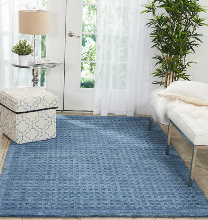 Blue textured area rug with a dotted square pattern in a bright living room; includes a patterned ottoman, glass side table, lamp, and plants.