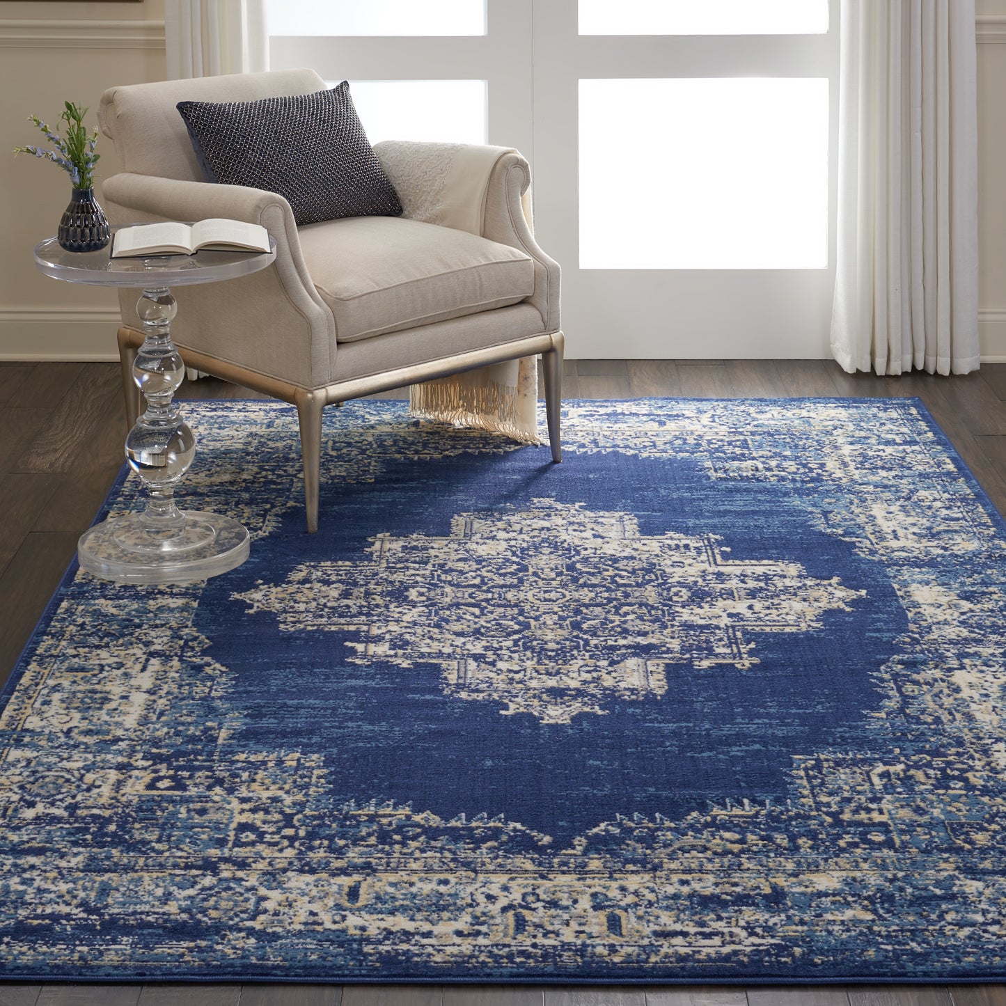 Beige fabric armchair with light wood legs beside a glass pedestal side table on a blue distressed oriental rug with ivory motifs.