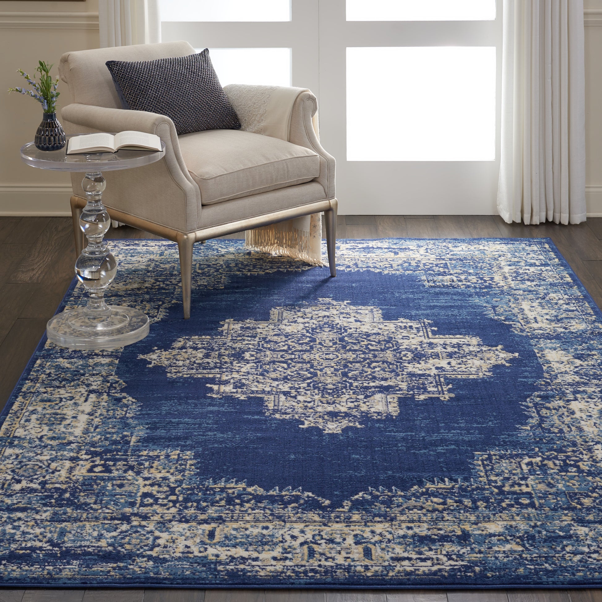 Beige fabric armchair with light wood legs beside a glass pedestal side table on a blue distressed oriental rug with ivory motifs.