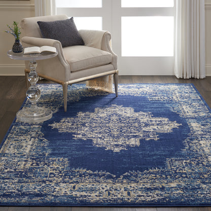 Beige fabric armchair with light wood legs beside a glass pedestal side table on a blue distressed oriental rug with ivory motifs.