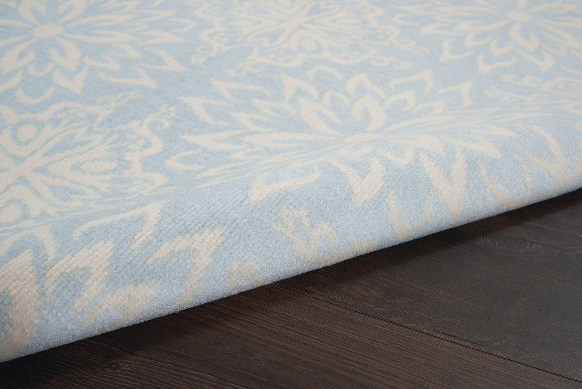 Light blue rug with cream damask floral pattern, folded edge on dark wooden floor.