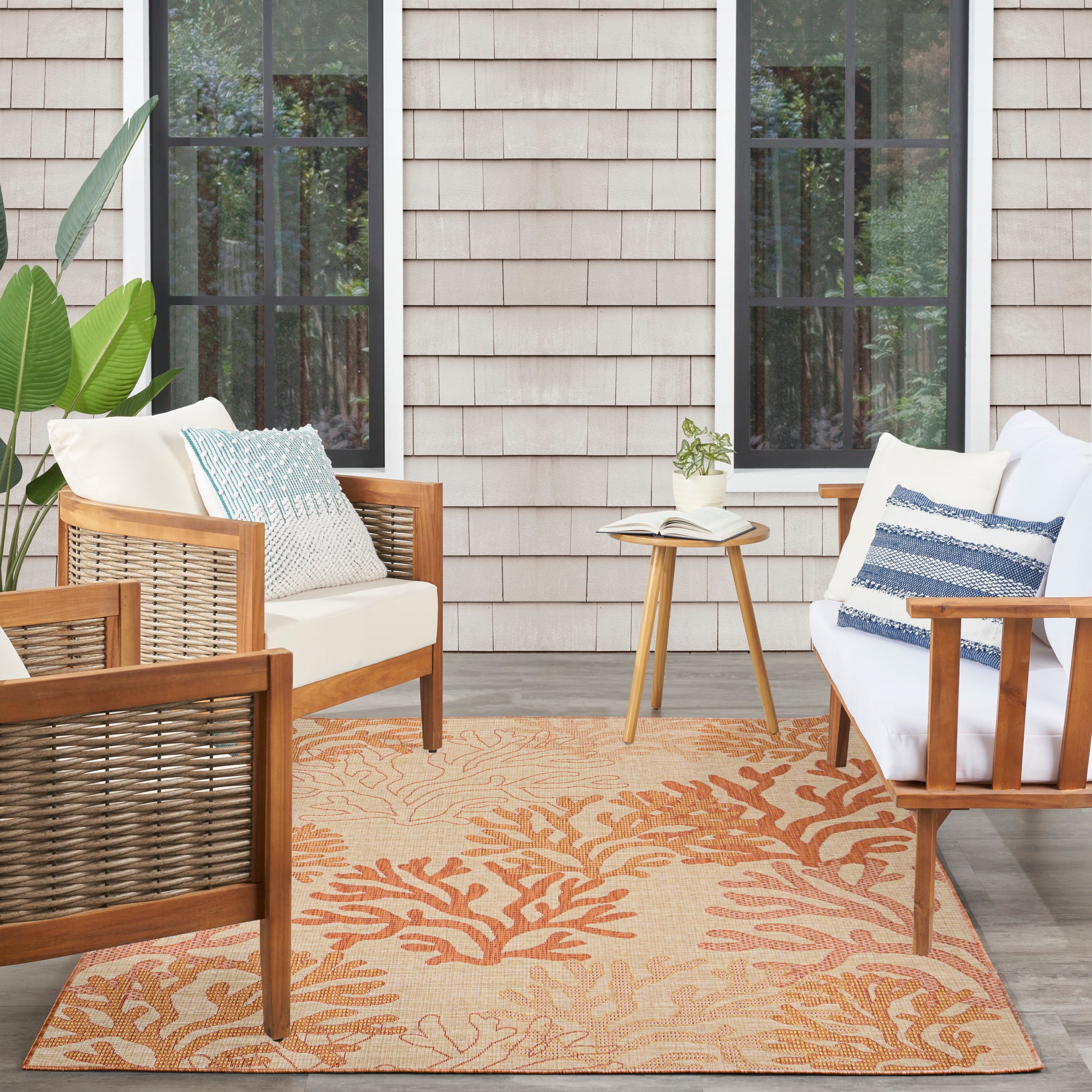 Outdoor seating with wooden wicker chairs, white cushions, blue pillows, small side table, plant, and orange coral rug.