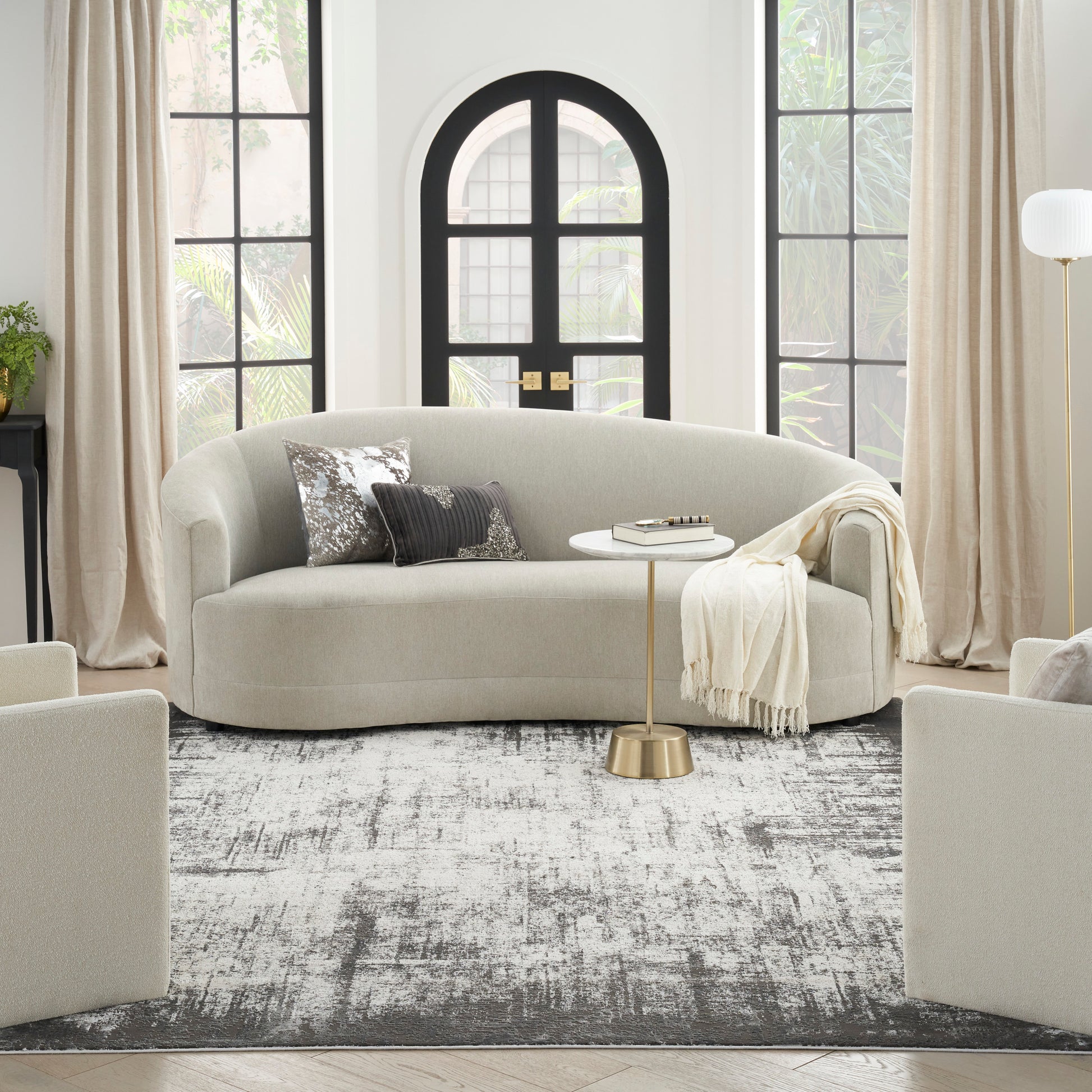Curved beige fabric sofa with silver sequin & charcoal pillows, round white-top gold-base side table, beige throw.