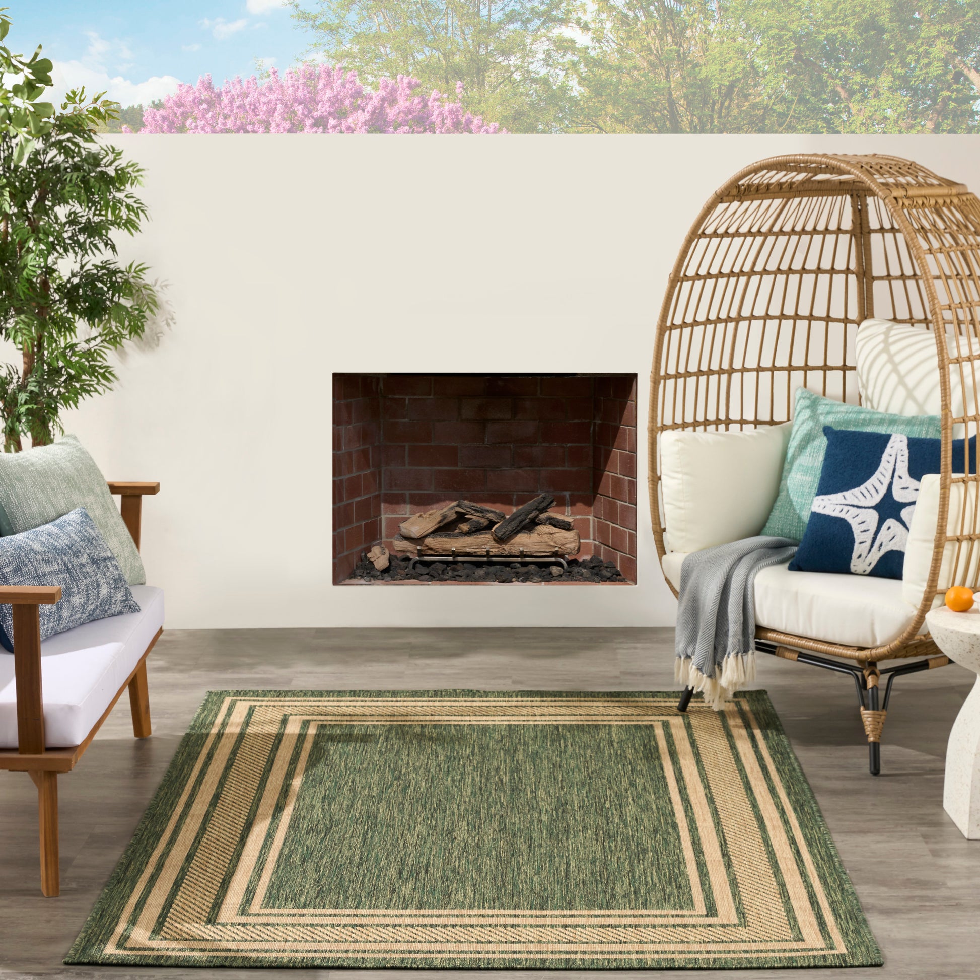 Outdoor patio with built-in brick fireplace, layered green rug, natural wicker egg chair with blue cushions, wooden seating.