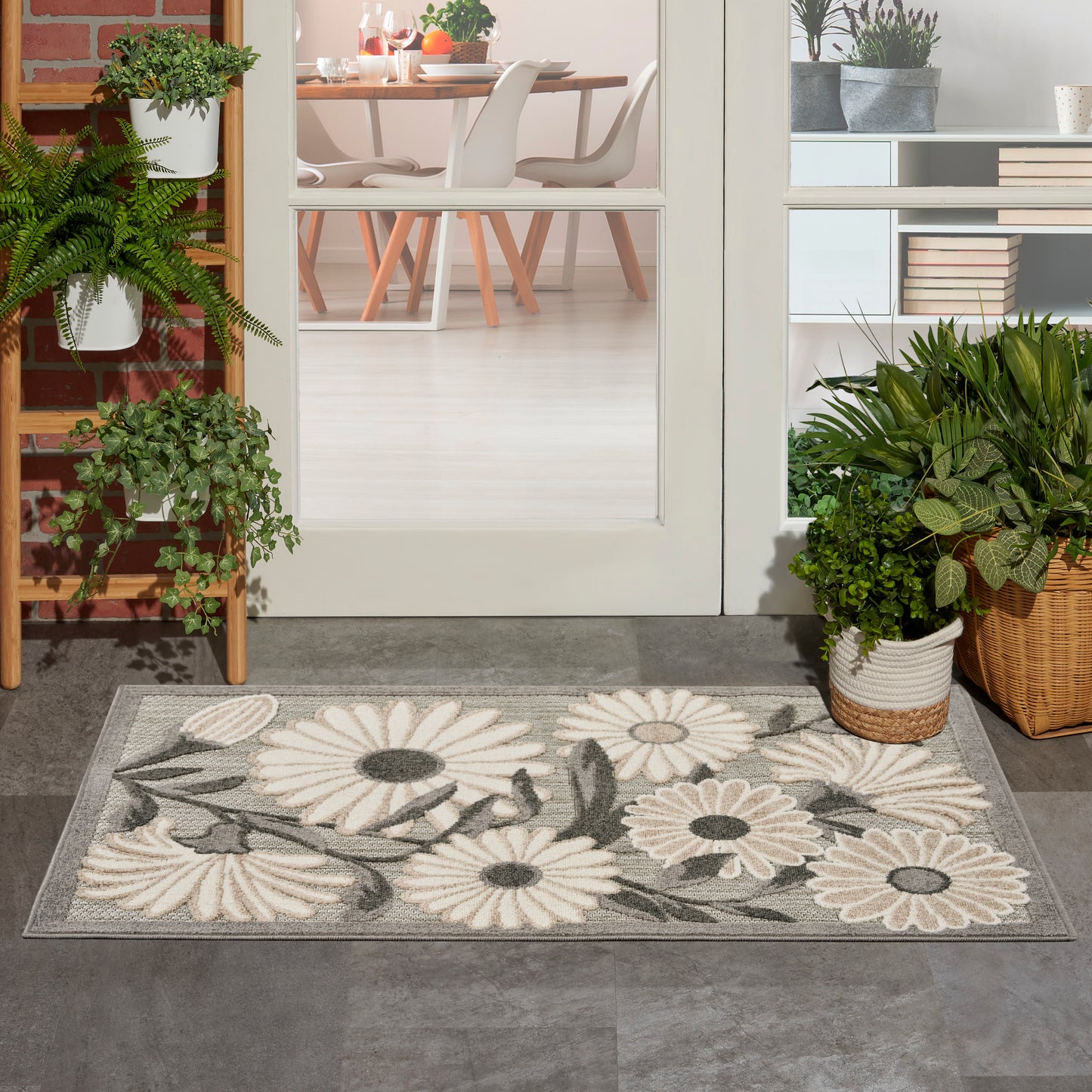 Floral rug with cream daisies on a gray background, 3' x 4', placed in an entry area with potted plants and a glass-door dining room visible in the background.