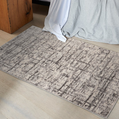 Rectangular gray-beige abstract area rug on light wood floor beside a bed with white and gray draped bedding.
