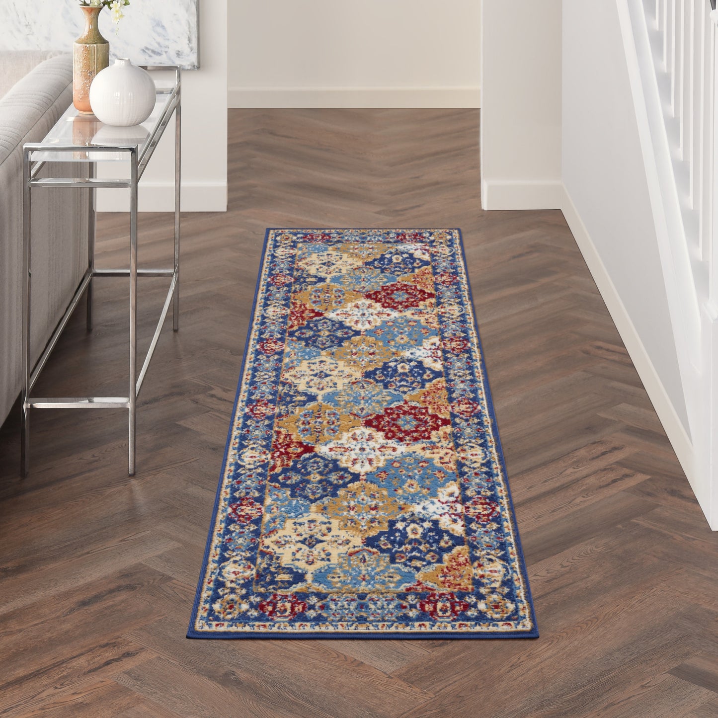 Runner rug with blue, red, ivory and gold geometric floral pattern in a narrow hallway with dark herringbone floor.