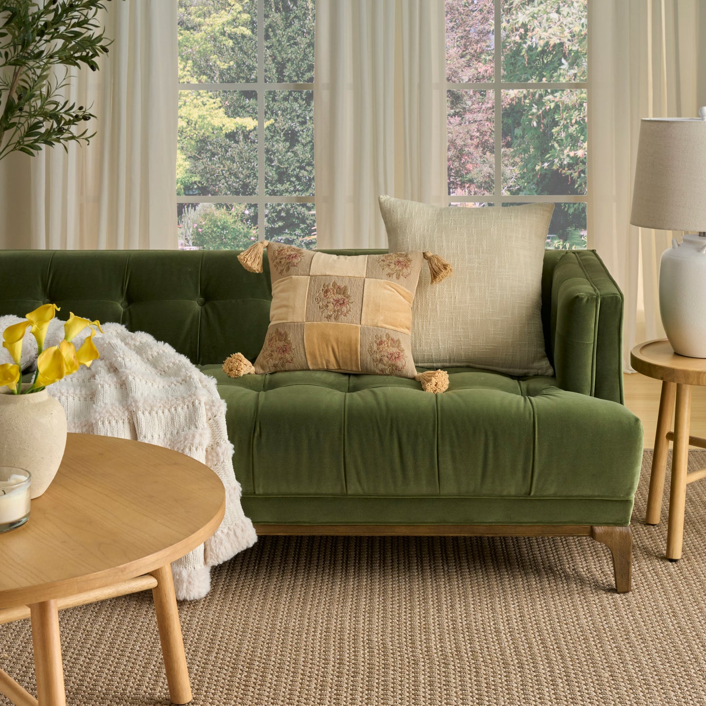 Green tufted sofa with beige cushions, cream throw, round wooden coffee table and yellow flowers in a bright living room.