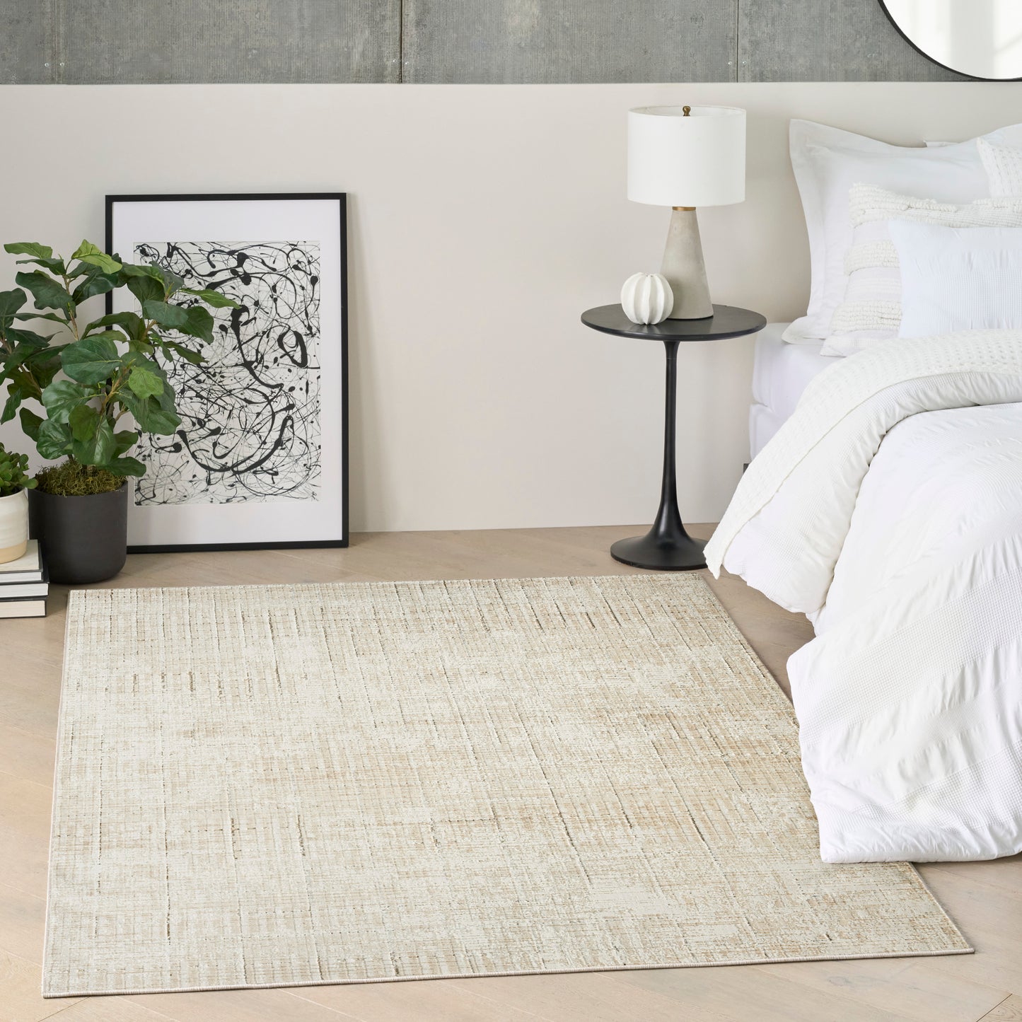 Neutral bedroom with beige woven rug, white bedding, round black pedestal nightstand, white lamp, abstract art, and greenery.