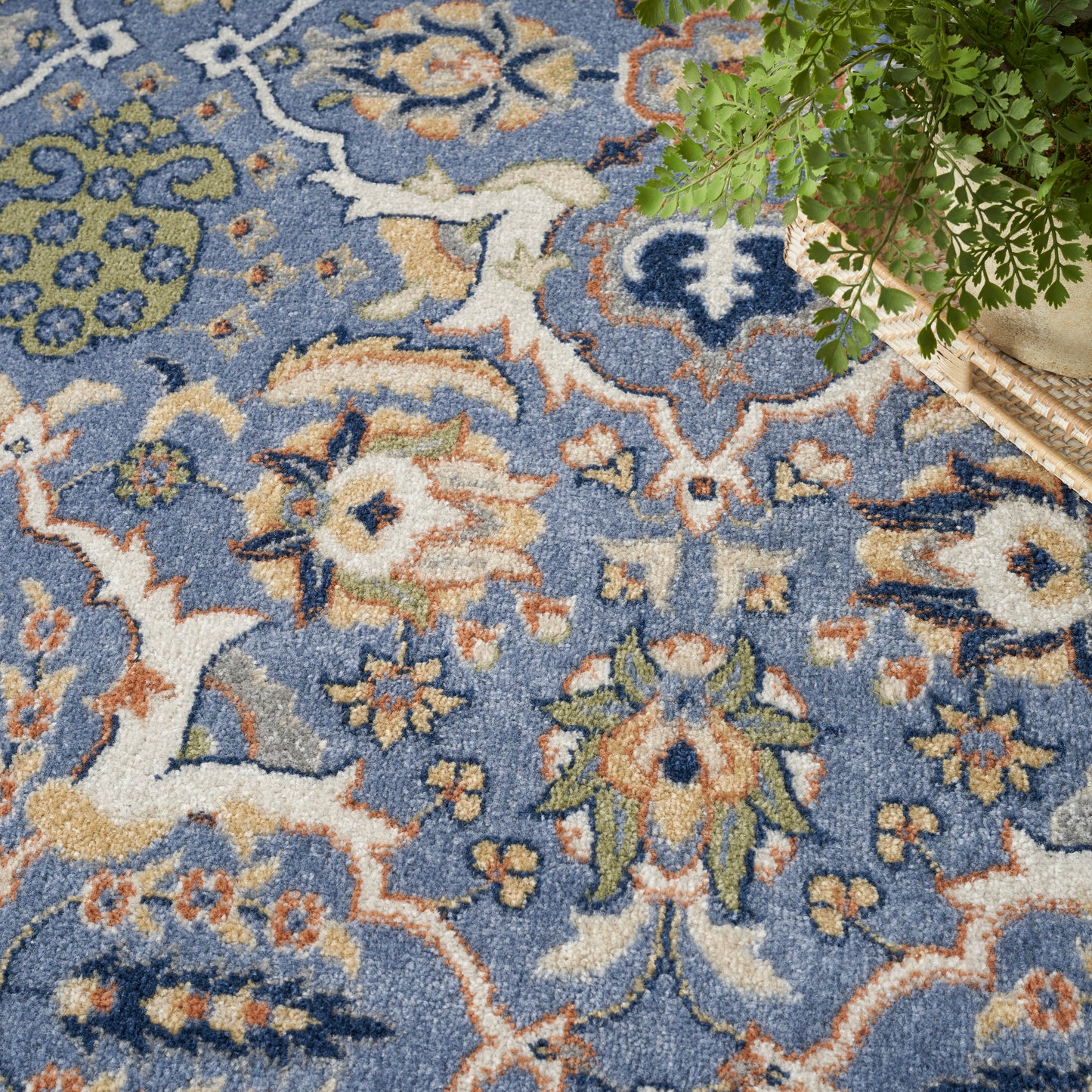 Blue bohemian rug with intricate cream, orange and green floral motifs; plant in wicker basket sits at corner.