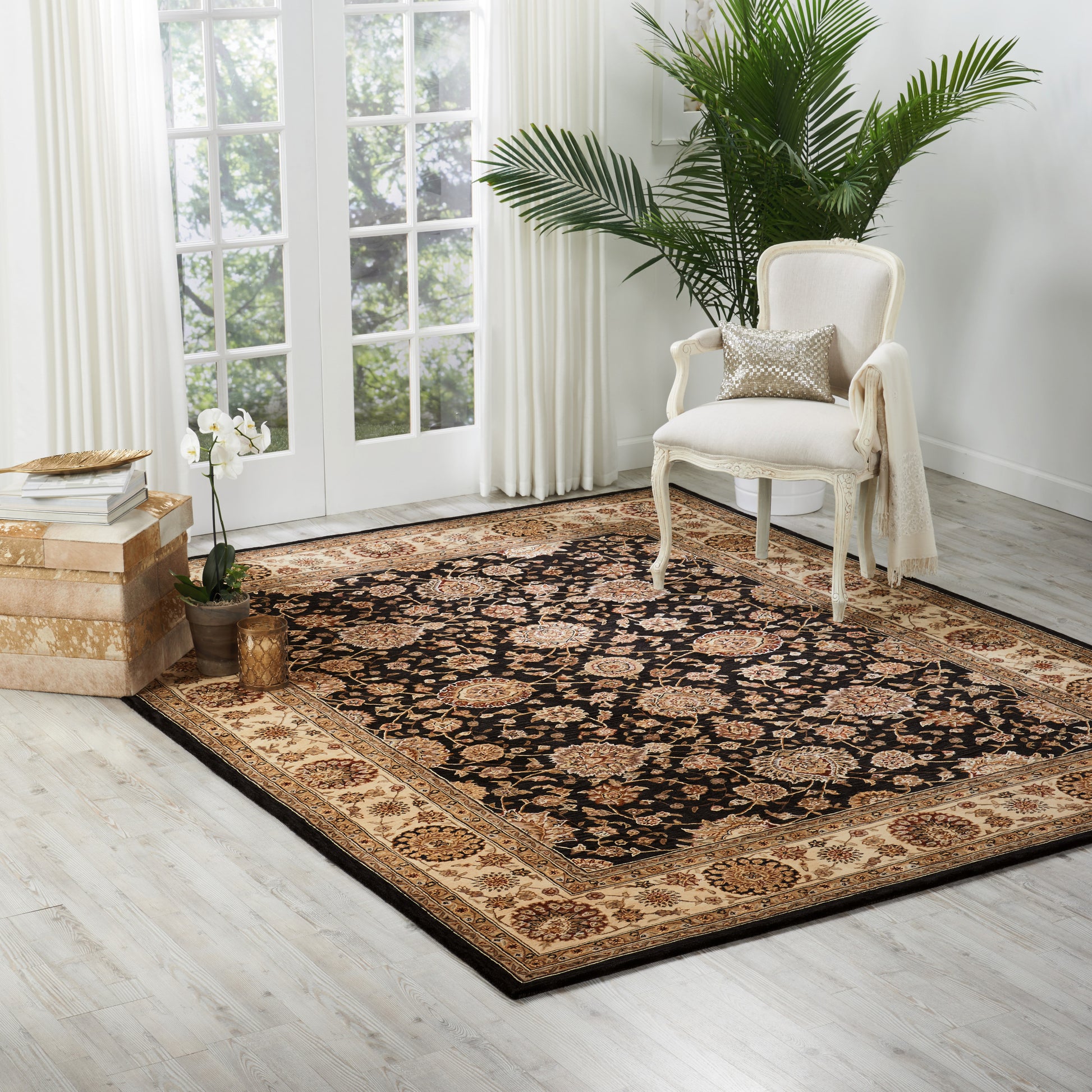 Persian-style area rug with black field and tan-gold border; white upholstered armchair, potted plants, light wood floor.