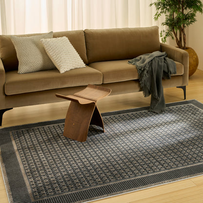 Taupe mid-century sofa with textured pillows, dark throw, curved wooden stool, geometric rug, and plant.