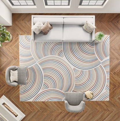 Rectangular area rug with overlapping concentric arc pattern in blue, beige and gray on a warm wood floor beneath a gray sofa.