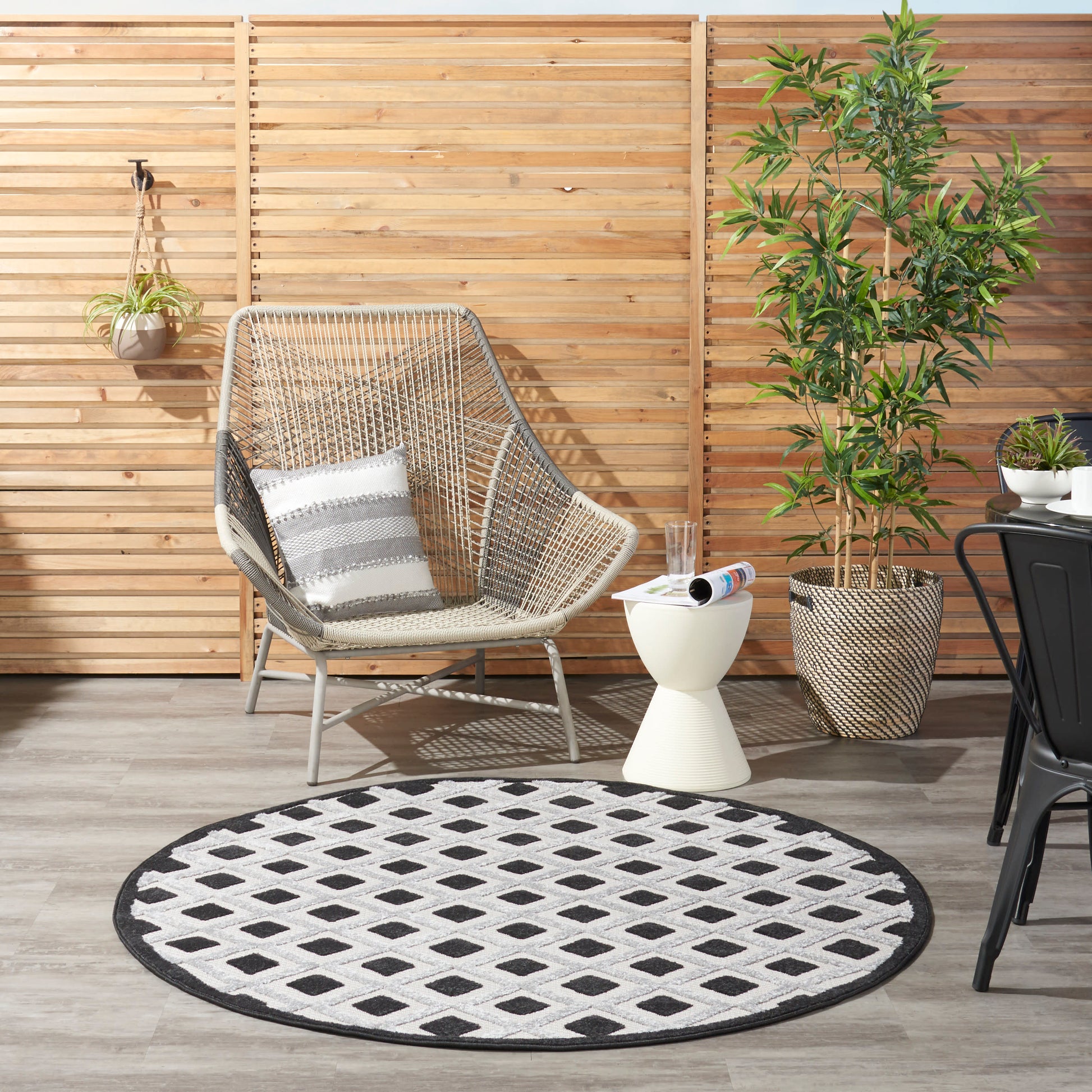 Outdoor wicker lounge chair with striped cushion, round black-and-white lattice rug, wooden privacy fence, tall potted plant.