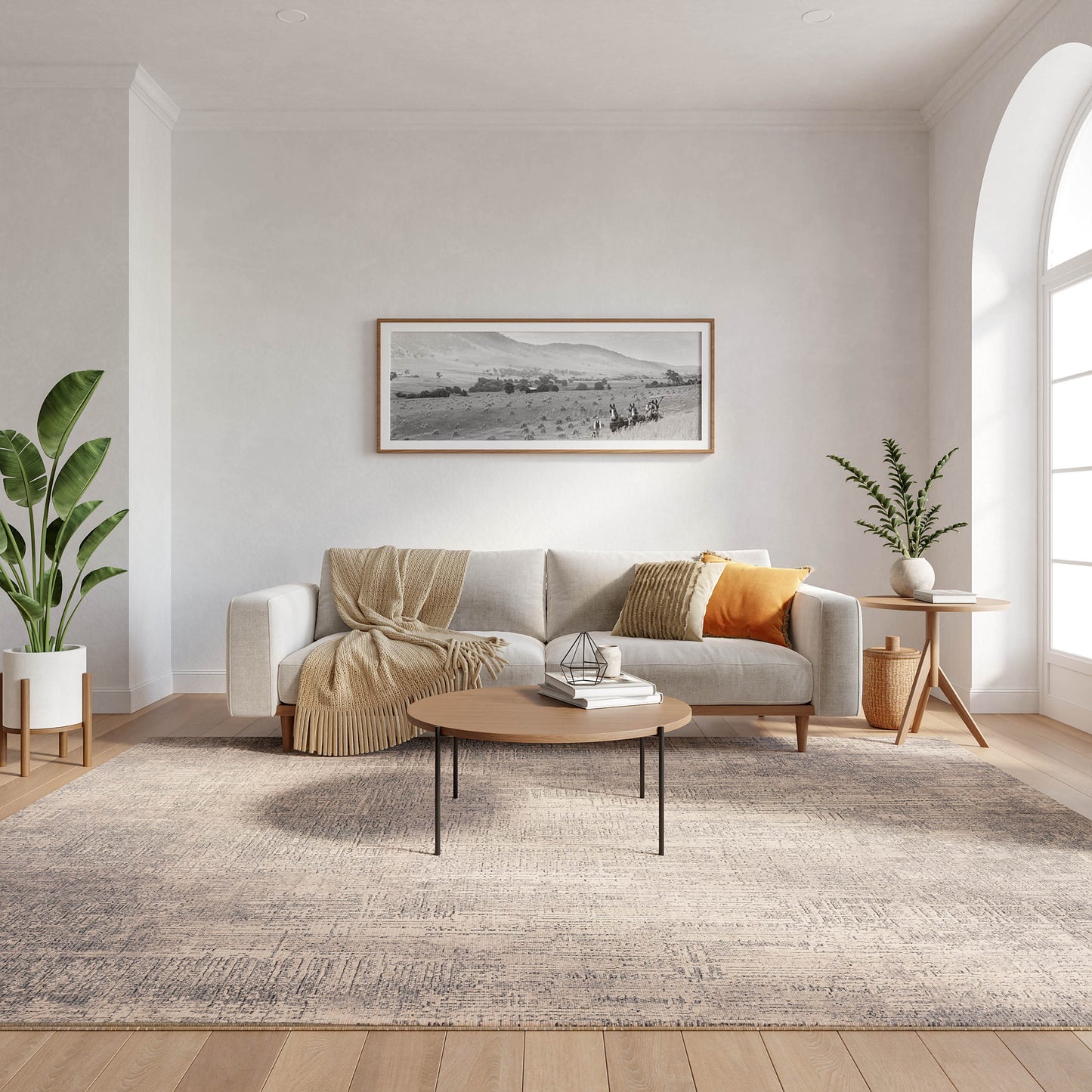 Neutral living room with light sofa, beige throw, orange cushion, framed landscape art, oval wooden coffee table, potted plants, arched window, grey rug.