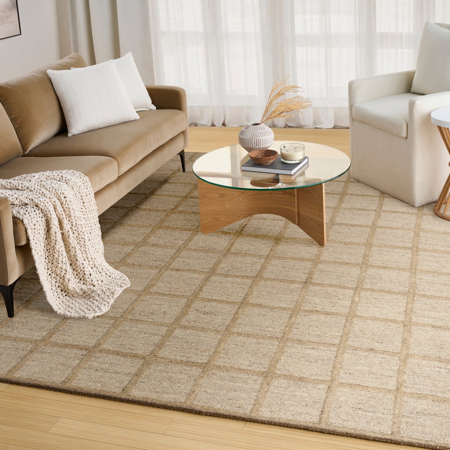 Neutral living room with beige sofa, white pillows, chunky knit throw, round glass-top coffee table and beige grid rug.