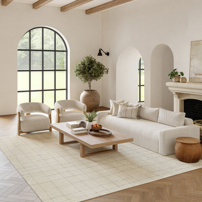 Neutral living room: cream sofa, curved chairs, wooden coffee table, beige rug, arched window, potted tree, fireplace.