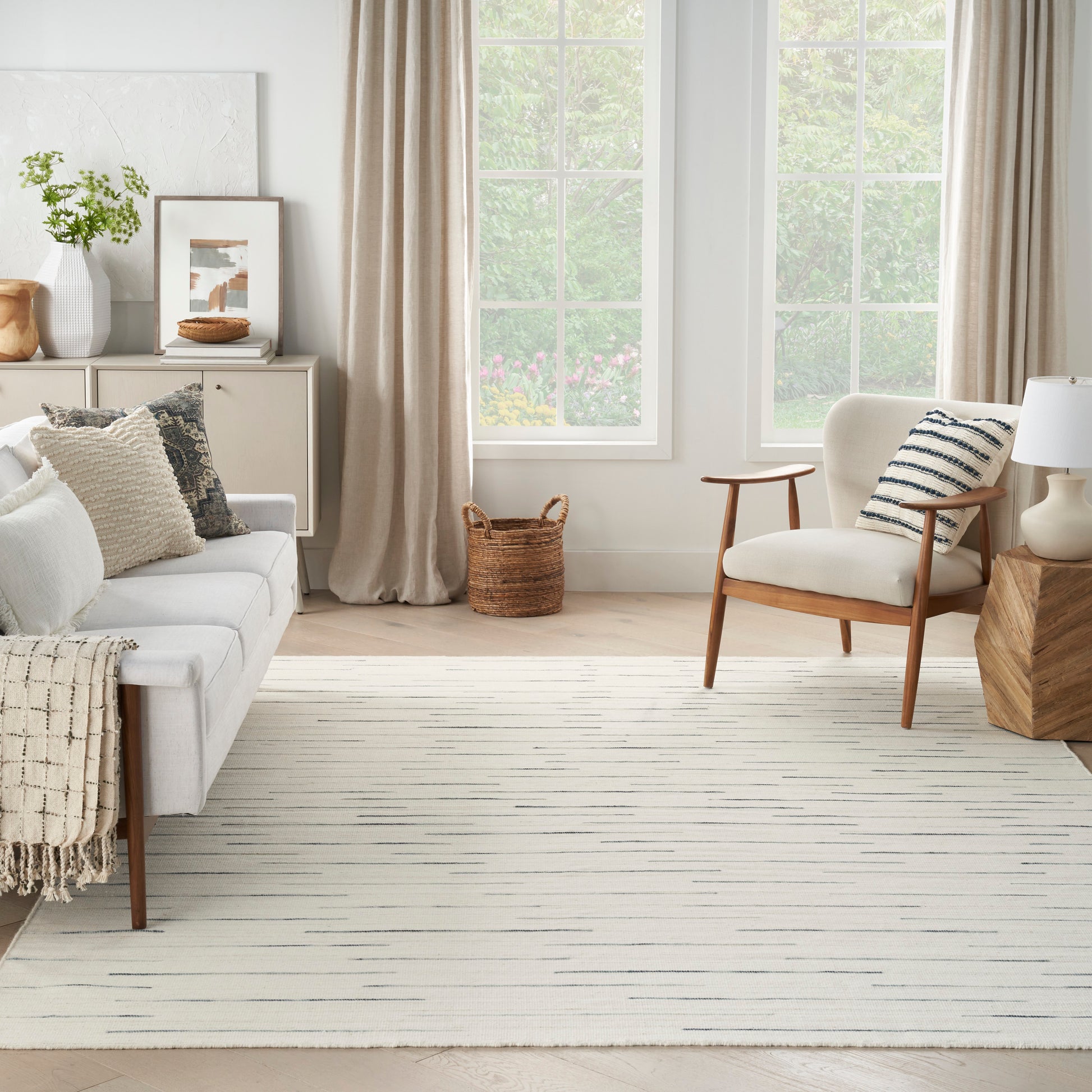 Neutral living room with cream sofa, mid-century wood chair, beige curtains, striped rug, woven basket, plant, framed art.
