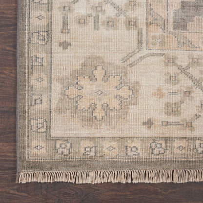 Beige patterned rug corner with tassel fringe, muted floral medallion design and bordered trim on dark wood floor.